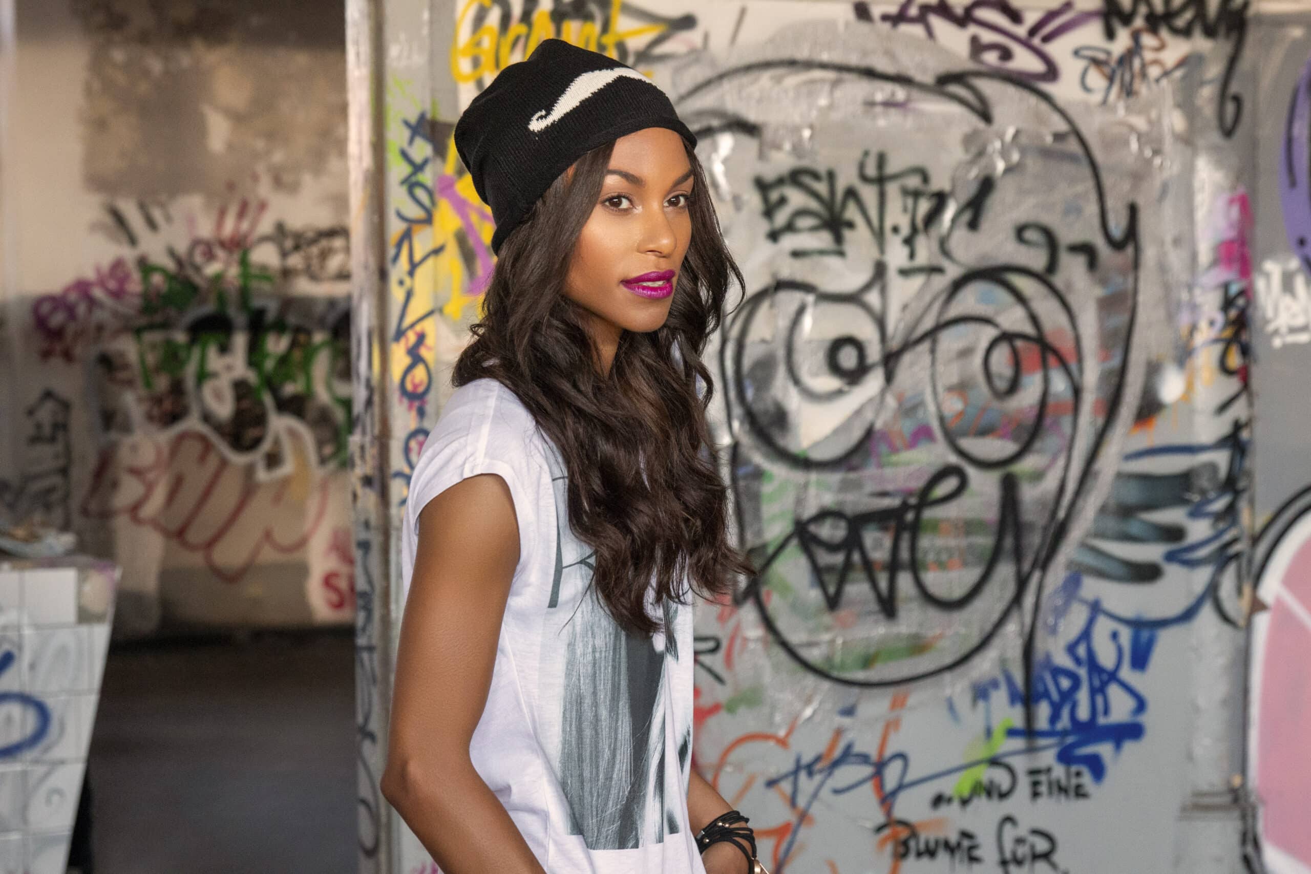 Eine selbstbewusste Frau mit langen, welligen Haaren, einer Beanie-Mütze und einem weißen T-Shirt steht vor einer mit Graffiti bedeckten Wand und blickt leicht zur Seite. © Fotografie Tomas Rodriguez