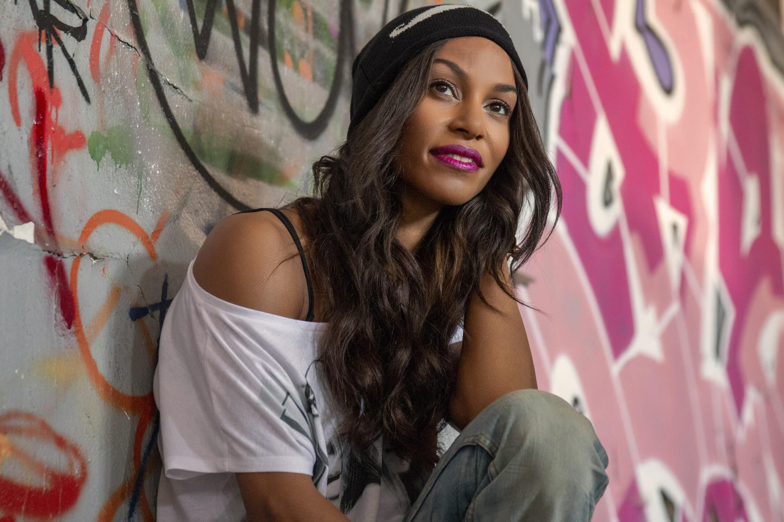 Eine Frau mit Beanie-Mütze sitzt lässig an einer mit Graffiti bedeckten Wand, trägt ein weißes T-Shirt und blickt mit einem leichten Lächeln zur Seite. © Fotografie Tomas Rodriguez