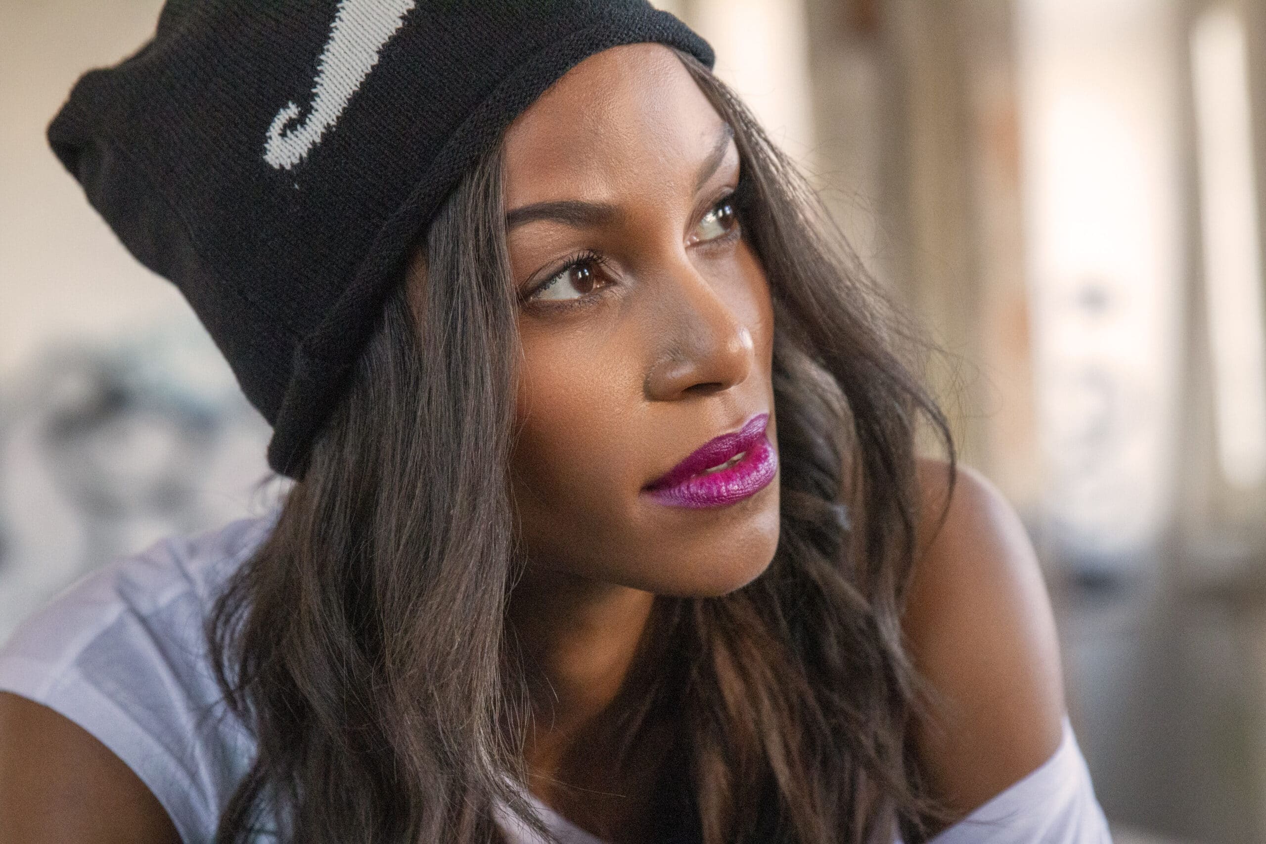 Eine Frau mit nachdenklichem Gesichtsausdruck trägt eine Beanie-Mütze und ein weißes T-Shirt. Ihr Make-up besteht aus auffälligem lila Lippenstift und sie blickt zur Seite. © Fotografie Tomas Rodriguez