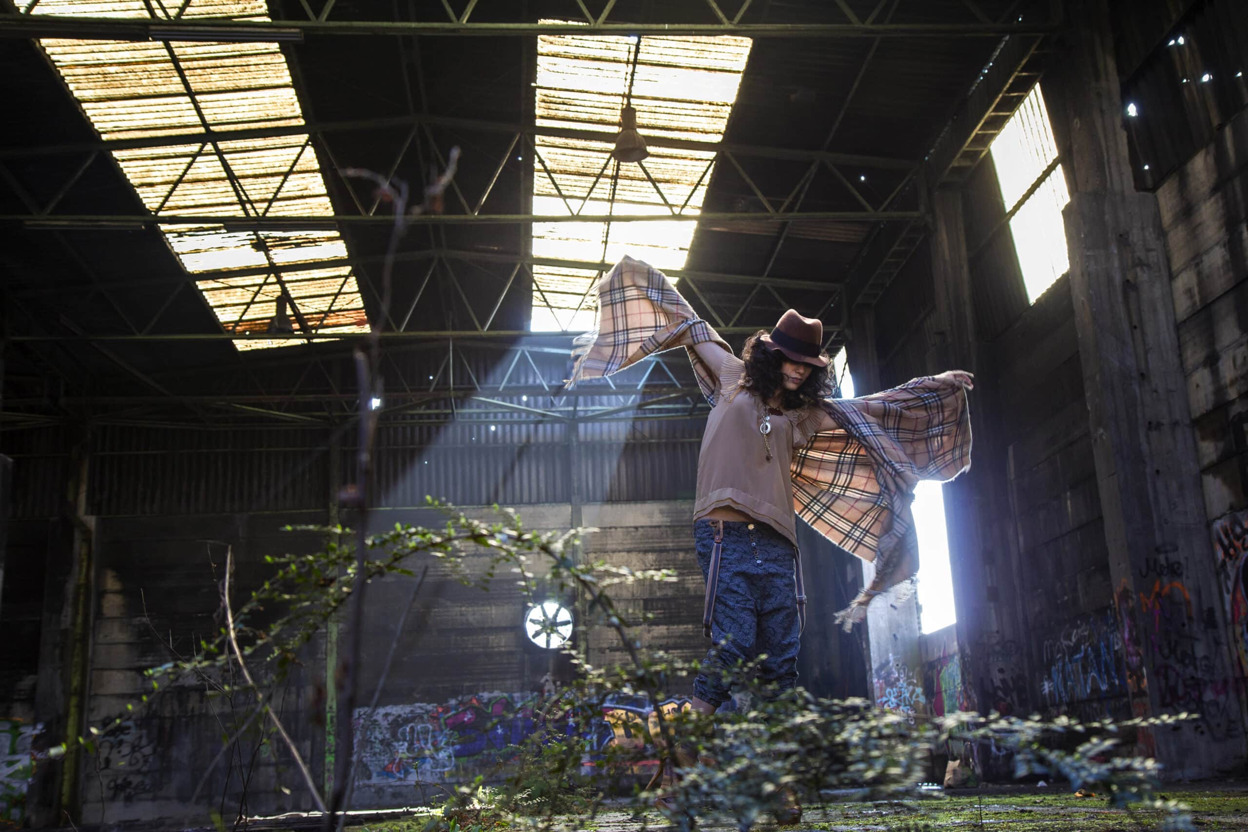 Eine Person mit Hut und Flanellhemd steht mit ausgebreiteten Armen in einem verlassenen, mit Graffiti bedeckten Lagerhaus. Sonnenlicht fällt durch zerbrochene Dachplatten, beleuchtet die Figur und erzeugt einen dramatischen Effekt. © Fotografie Tomas Rodriguez