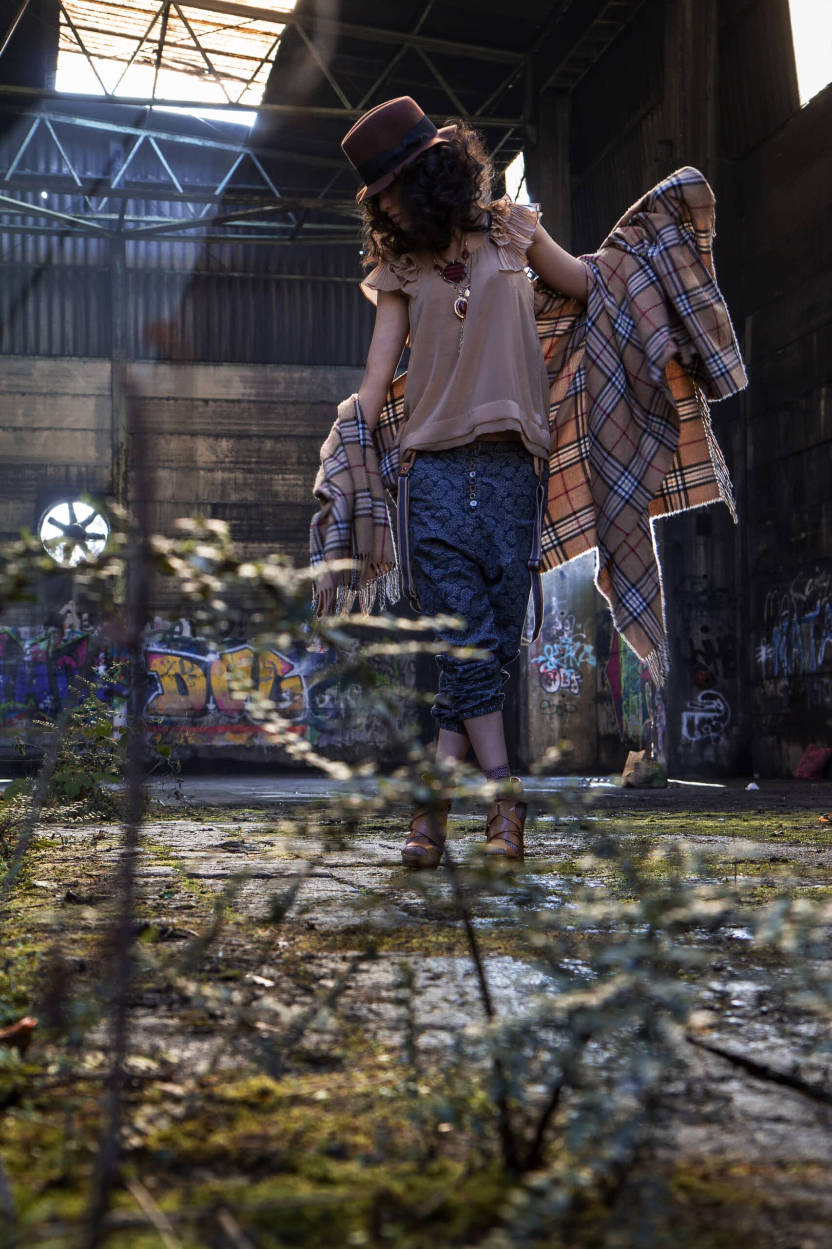 Eine Frau in einem stylischen Outfit, darunter Hut und wallender Schal, dreht sich fröhlich in einer mit Graffiti besprühten, sonnendurchfluteten Lagerhalle, umgeben von Grün. © Fotografie Tomas Rodriguez