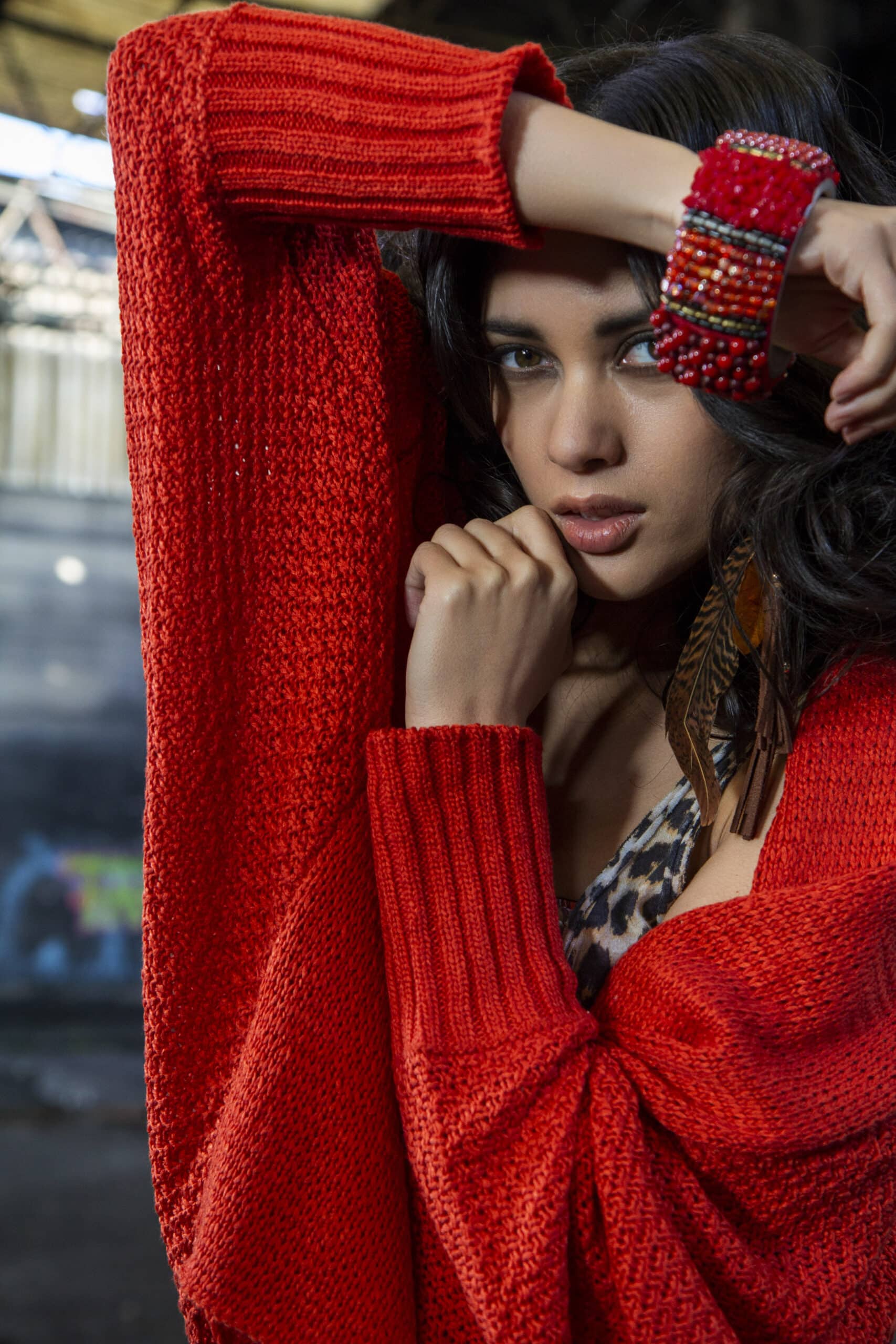 Eine Frau in einem leuchtend roten Pullover posiert mit erhobenem Arm und präsentiert ein buntes Armband. Ihr Look wird durch Federohrringe und einen Blick auf ein Oberteil mit Leopardenmuster akzentuiert. © Fotografie Tomas Rodriguez