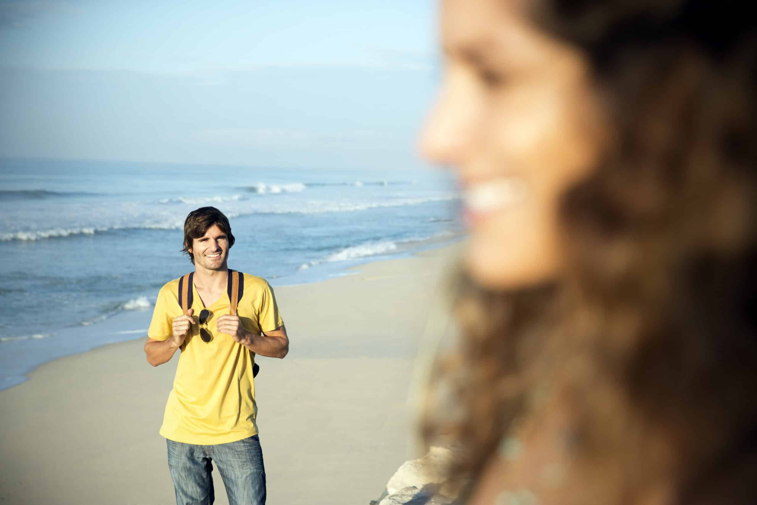 Ein Mann mit Rucksack und gelbem Hemd geht am Strand entlang und lächelt im Hintergrund. Im unscharfen Vordergrund lächelt eine Frau mit lockigem Haar und blickt in die Ferne. Am Ufer sind das Meer und die Wellen zu sehen. © Fotografie Tomas Rodriguez