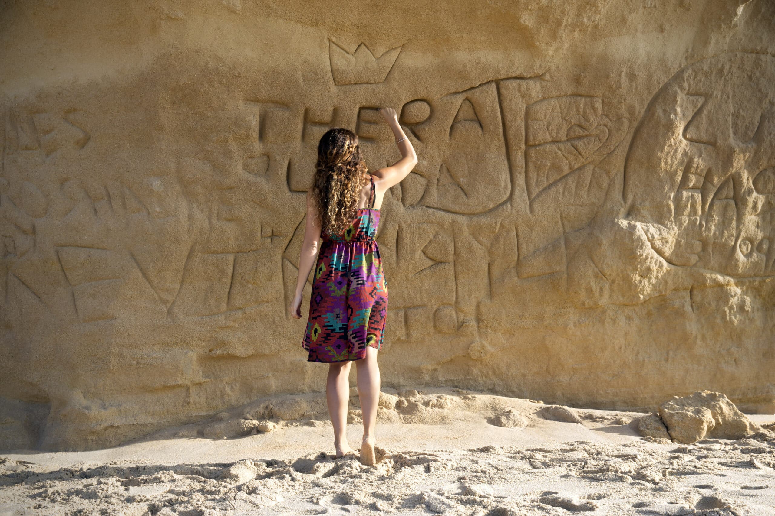 Eine Frau mit lockigem Haar und einem bunten Kleid steht an einem Sandstrand vor einer Felswand. Sie ritzt oder zeichnet mit ihrem Finger Wörter und Symbole in die Felsoberfläche. Die Wand weist verschiedene Gravuren auf, darunter eine Krone, Buchstaben und Formen. © Fotografie Tomas Rodriguez