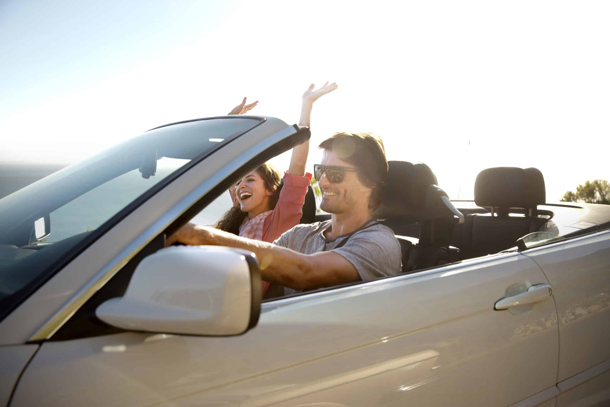 Ein lächelnder Mann fährt ein weißes Cabrio mit offenem Verdeck. Eine Frau auf dem Beifahrersitz hat ihre Hände freudig erhoben. Beide scheinen einen sonnigen Tag zu genießen, mit einem klaren und hellen Himmel im Hintergrund. © Fotografie Tomas Rodriguez