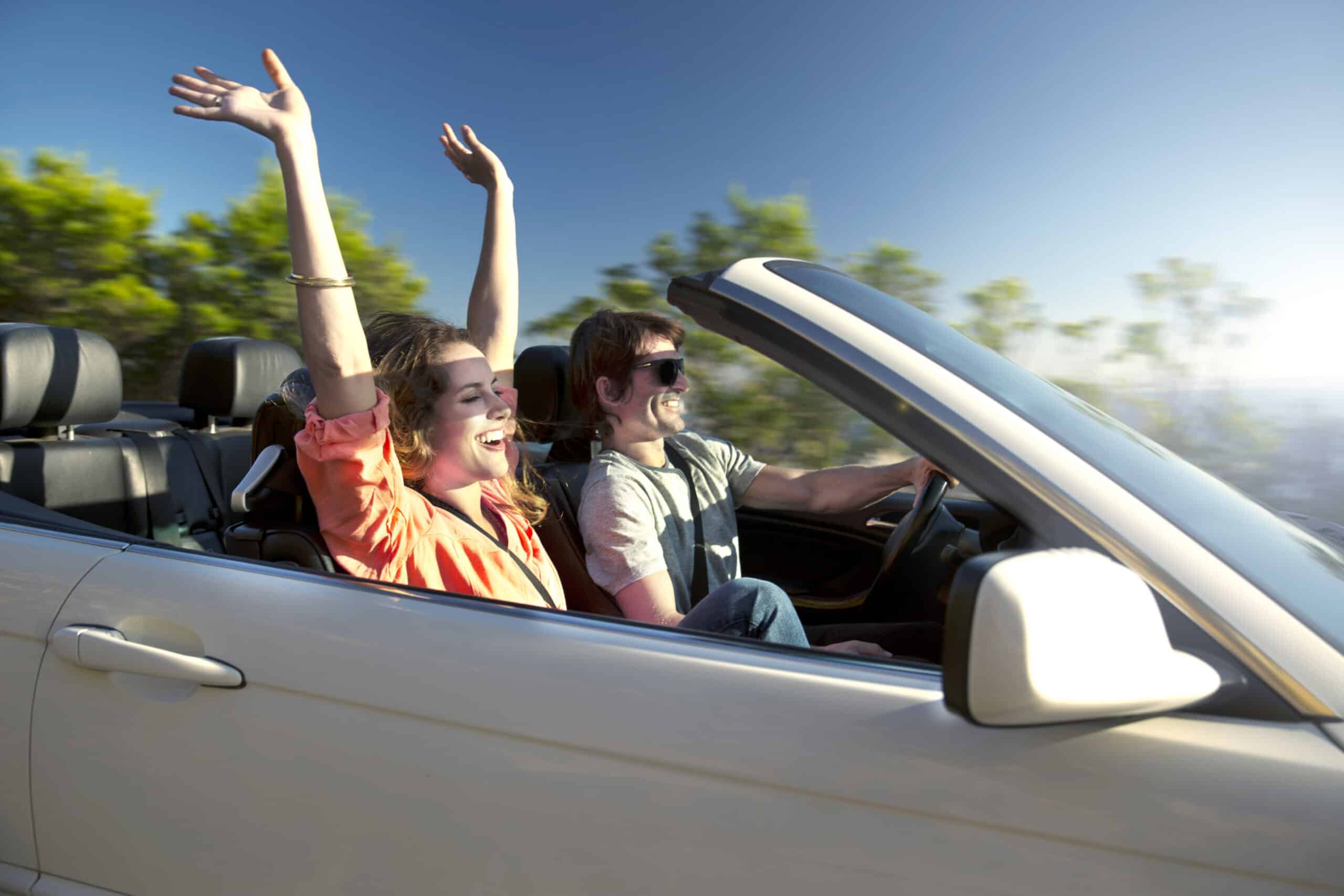 Zwei Personen genießen eine Fahrt in einem weißen Cabrio an einem sonnigen Tag. Der Beifahrer trägt eine orangefarbene Jacke mit erhobenen Armen und der Fahrer lächelt mit Sonnenbrille. Im Hintergrund ist ein verschwommenes Grün zu sehen, das auf Bewegung hindeutet. © Fotografie Tomas Rodriguez