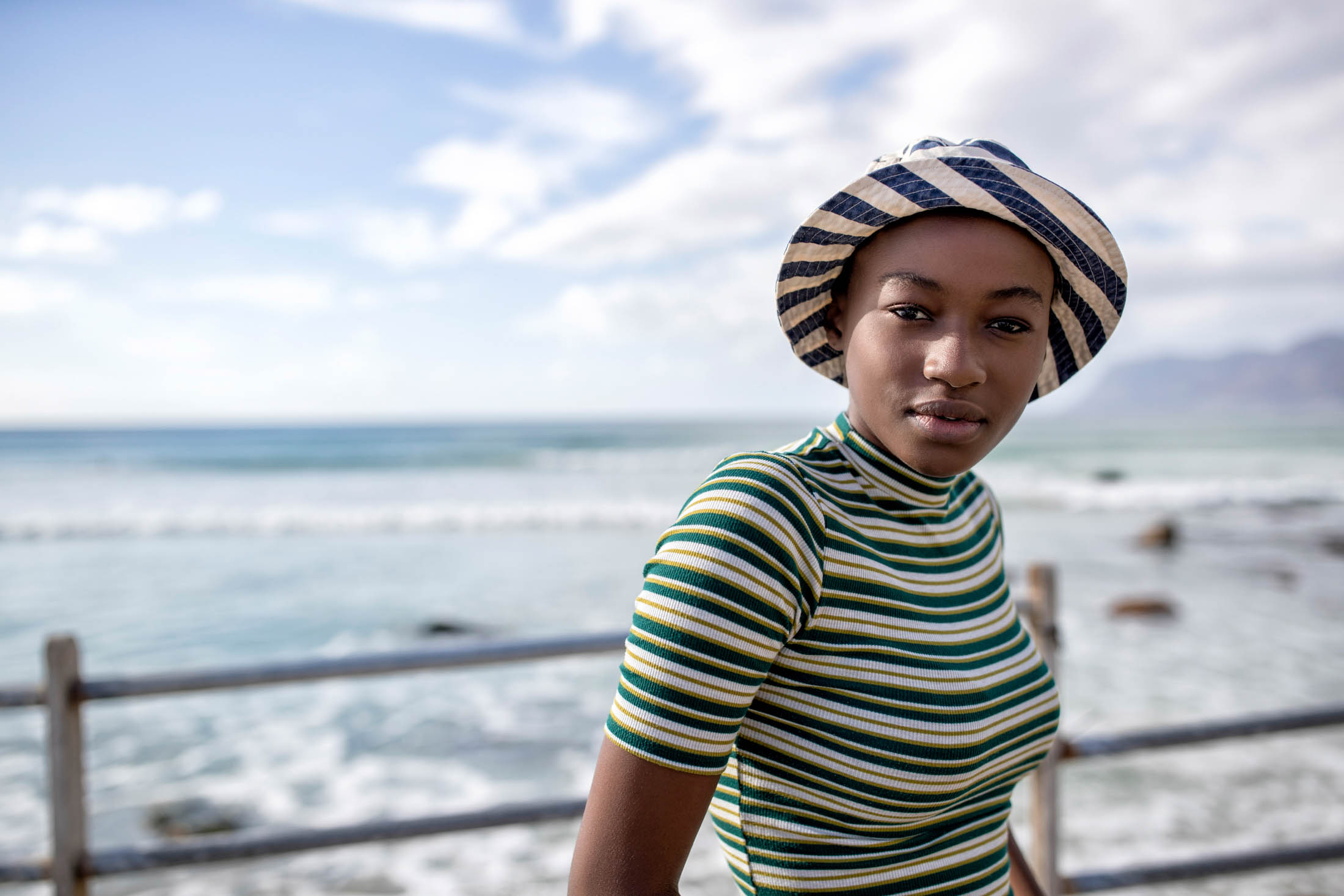 Eine junge Frau mit gestreiftem Hut und Oberteil steht an einem Geländer am Meer mit dem Meer und den Bergen in der Ferne im Hintergrund. Sie blickt direkt in die Kamera. © Fotografie Tomas Rodriguez