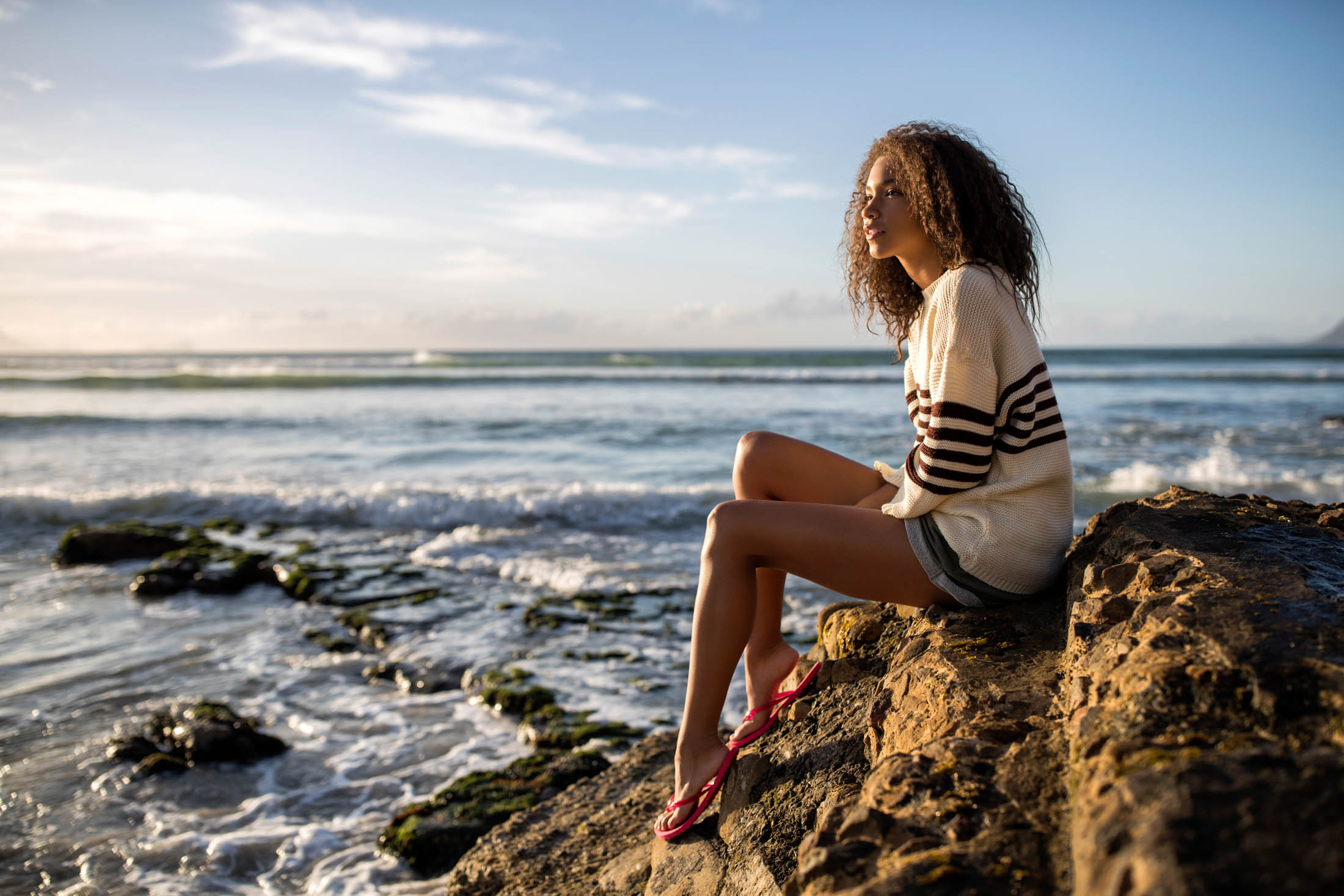 Eine Frau sitzt an einer felsigen Küste, blickt auf das Meer. Sie trägt einen gestreiften Pullover und rote Sandalen und ihr lockiges Haar wird sanft von der Meeresbrise zerzaust. © Fotografie Tomas Rodriguez