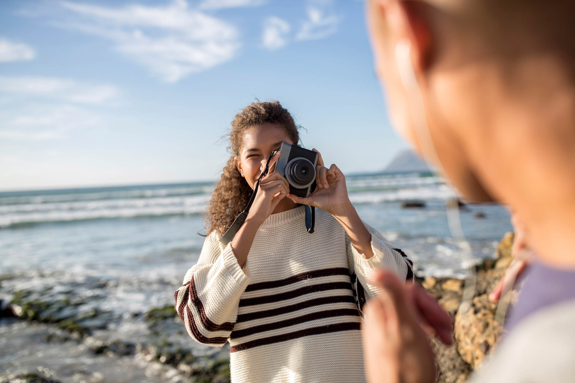 Eine junge Frau mit lockigem Haar macht mit einer DSLR-Kamera ein Foto von einer anderen Person am Meer. Sie befinden sich beide an einem felsigen Strand mit klarem Himmel und ruhigem Wasser im Hintergrund. © Fotografie Tomas Rodriguez