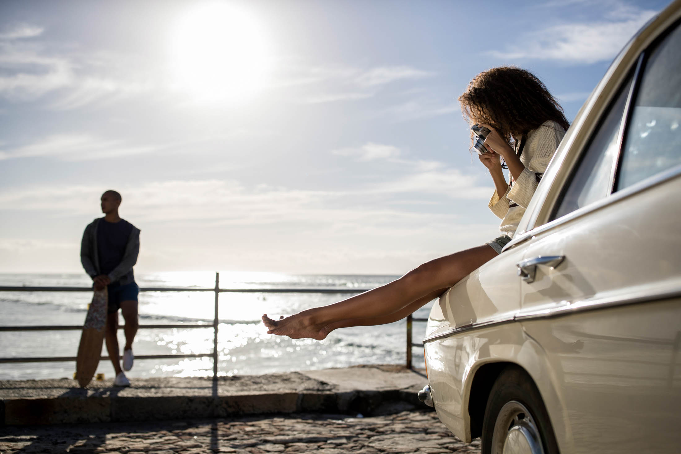 Eine Frau sitzt in einem Auto am Meer und streckt entspannt ihr Bein aus der Autotür, eine Kamera in der Hand. Im Hintergrund steht ein Mann mit Blick auf das Meer. © Fotografie Tomas Rodriguez