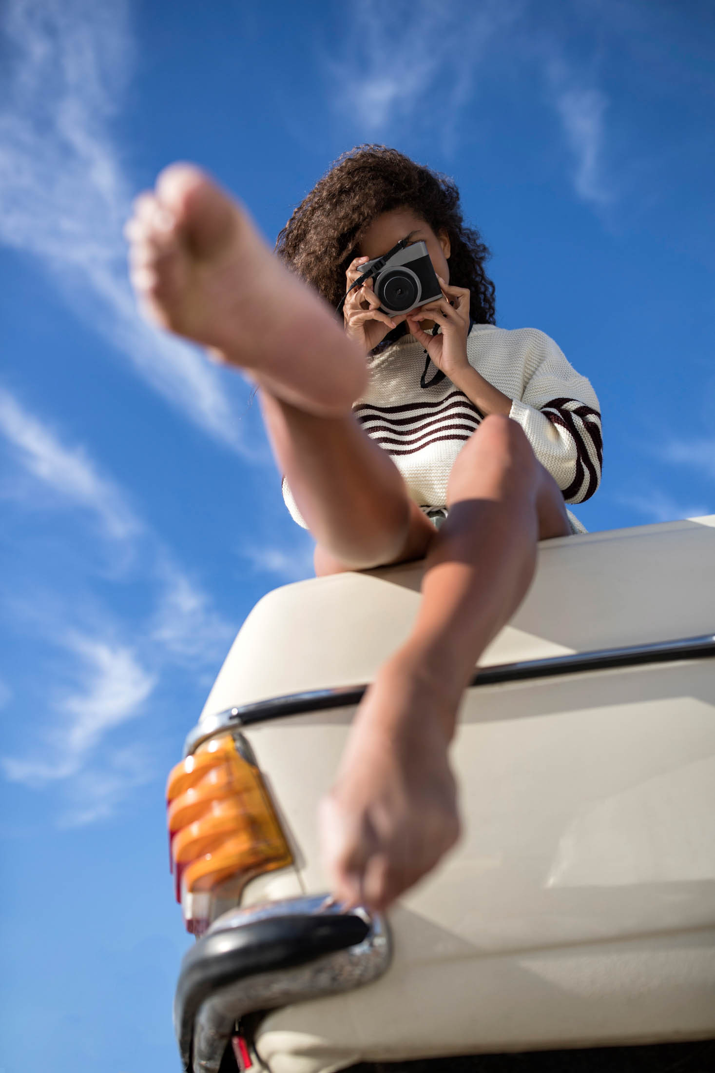 Eine Person mit lockigem Haar sitzt auf der Motorhaube eines Oldtimers und macht mit einer Kamera ein Foto. Ihr nackter Fuß ist im Vordergrund deutlich vor einem klaren blauen Himmel zu erkennen. © Fotografie Tomas Rodriguez