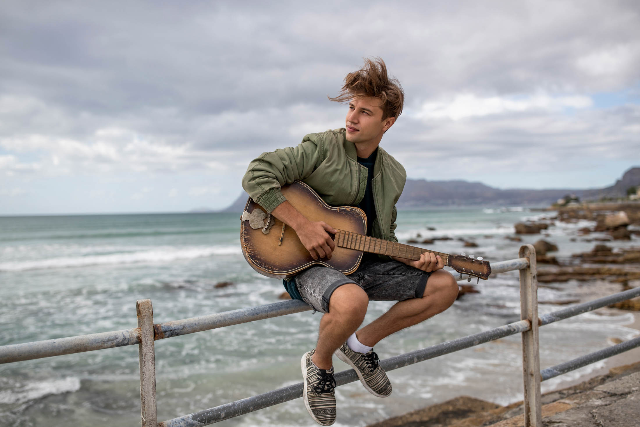 Ein junger Mann mit zerzaustem Haar sitzt auf einem Metallgeländer am Meeresufer und spielt Akustikgitarre. Berge und bewölkter Himmel bilden die Kulisse. © Fotografie Tomas Rodriguez