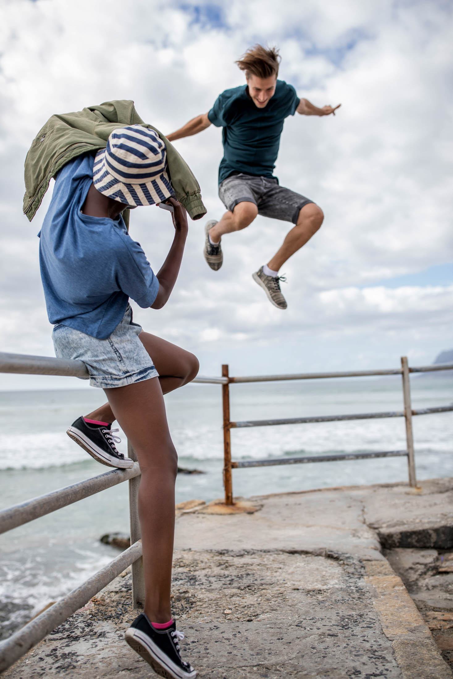 Ein kleiner Junge springt in der Luft von einem Geländer am Meer und wird dabei von einem anderen Jungen beobachtet, der seine Augen mit einem Hut abschirmt. Sie befinden sich auf einer Strandpromenade unter einem bewölkten Himmel. © Fotografie Tomas Rodriguez