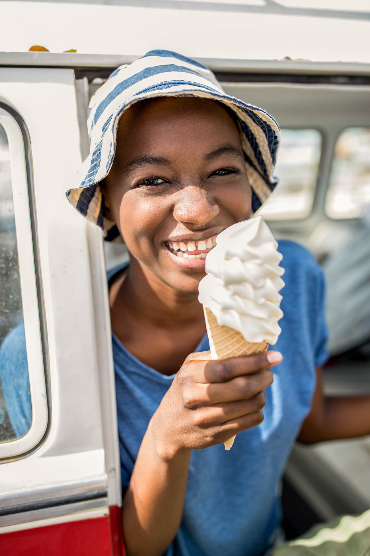 Eine fröhliche junge Frau mit gestreifter Mütze und blauem T-Shirt genießt an einem sonnigen Tag neben einem Lieferwagen eine große Vanilleeistüte. © Fotografie Tomas Rodriguez