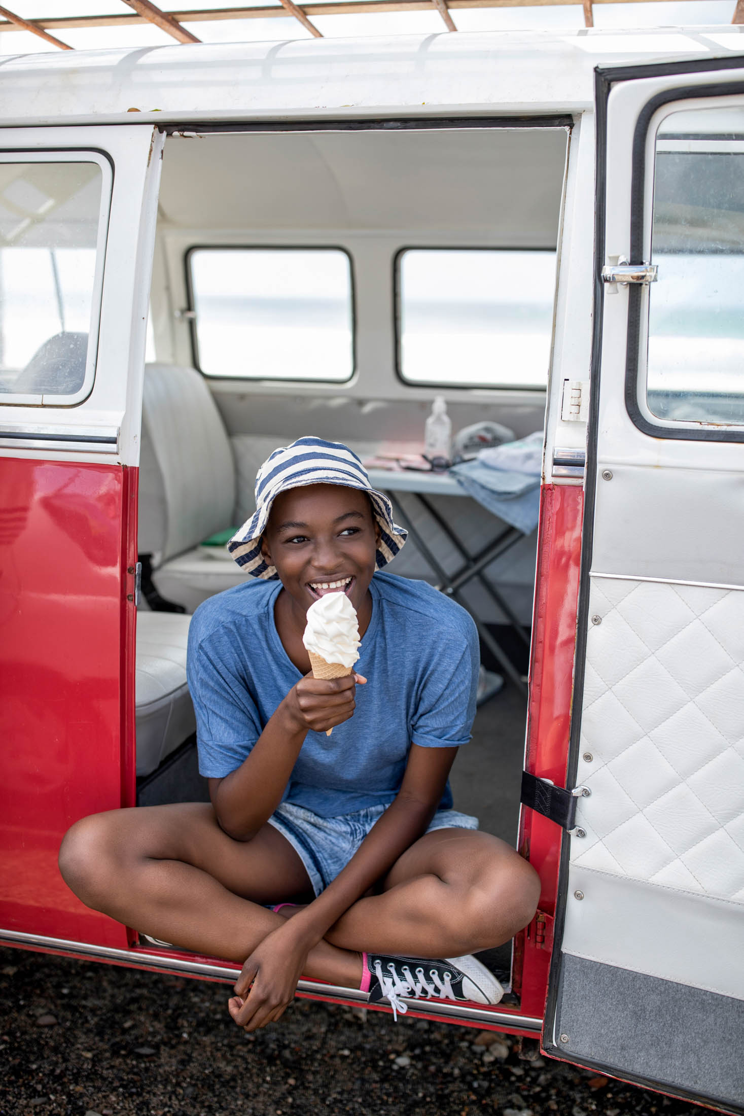 Eine junge Frau in einem blauen Kleid und einem gestreiften Hut sitzt in der Tür eines rot-weißen Oldtimer-Wohnmobils und lächelt, während sie eine Eistüte isst. © Fotografie Tomas Rodriguez