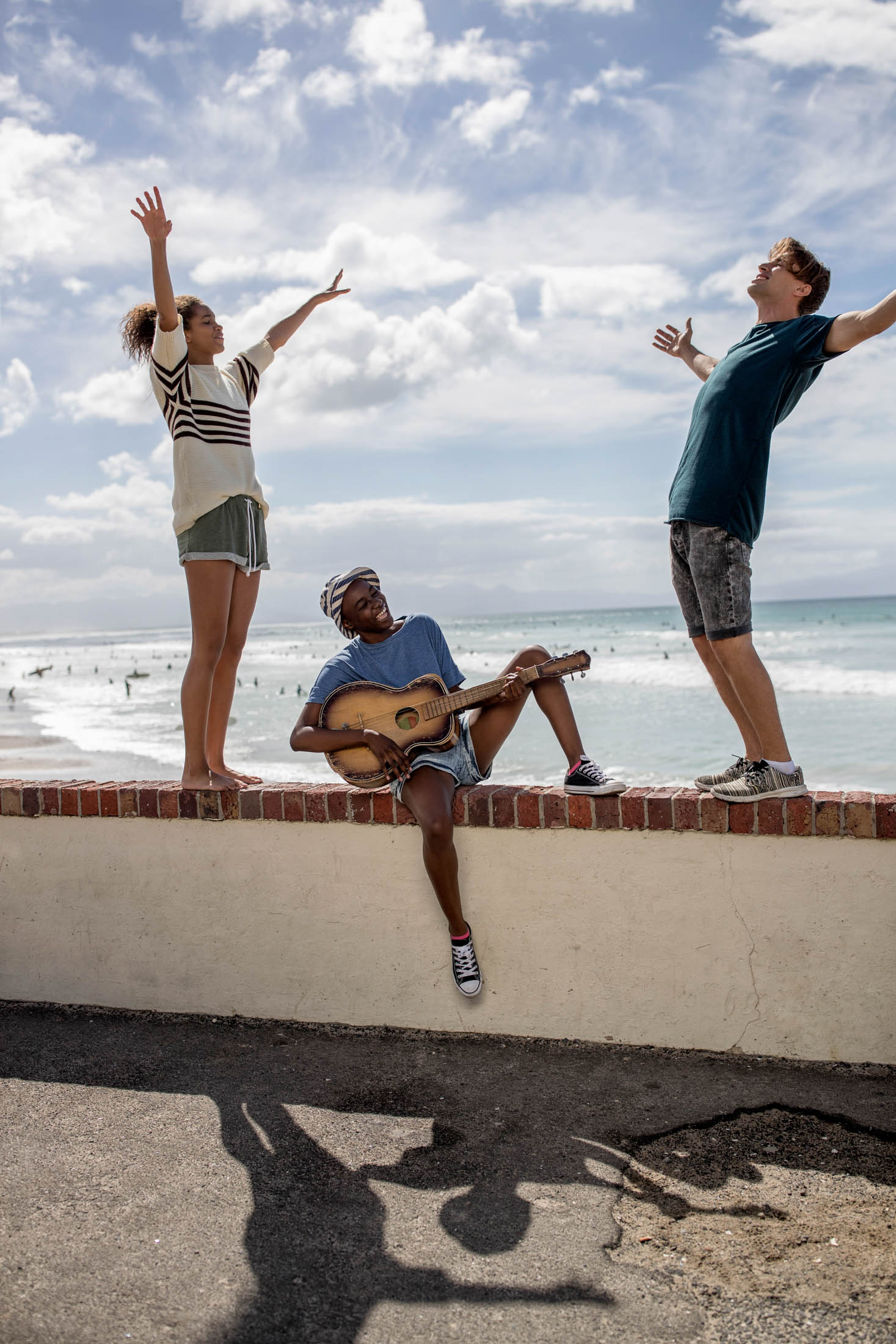 Drei Freunde genießen einen sonnigen Tag am Strand. Eine Person sitzt auf einer Mauer und spielt Gitarre, während zwei andere freudig ihre Arme gen Himmel strecken und so ihre unbeschwerte Aufregung ausdrücken. Das Meer voller Surfer bietet eine ruhige Kulisse. © Fotografie Tomas Rodriguez