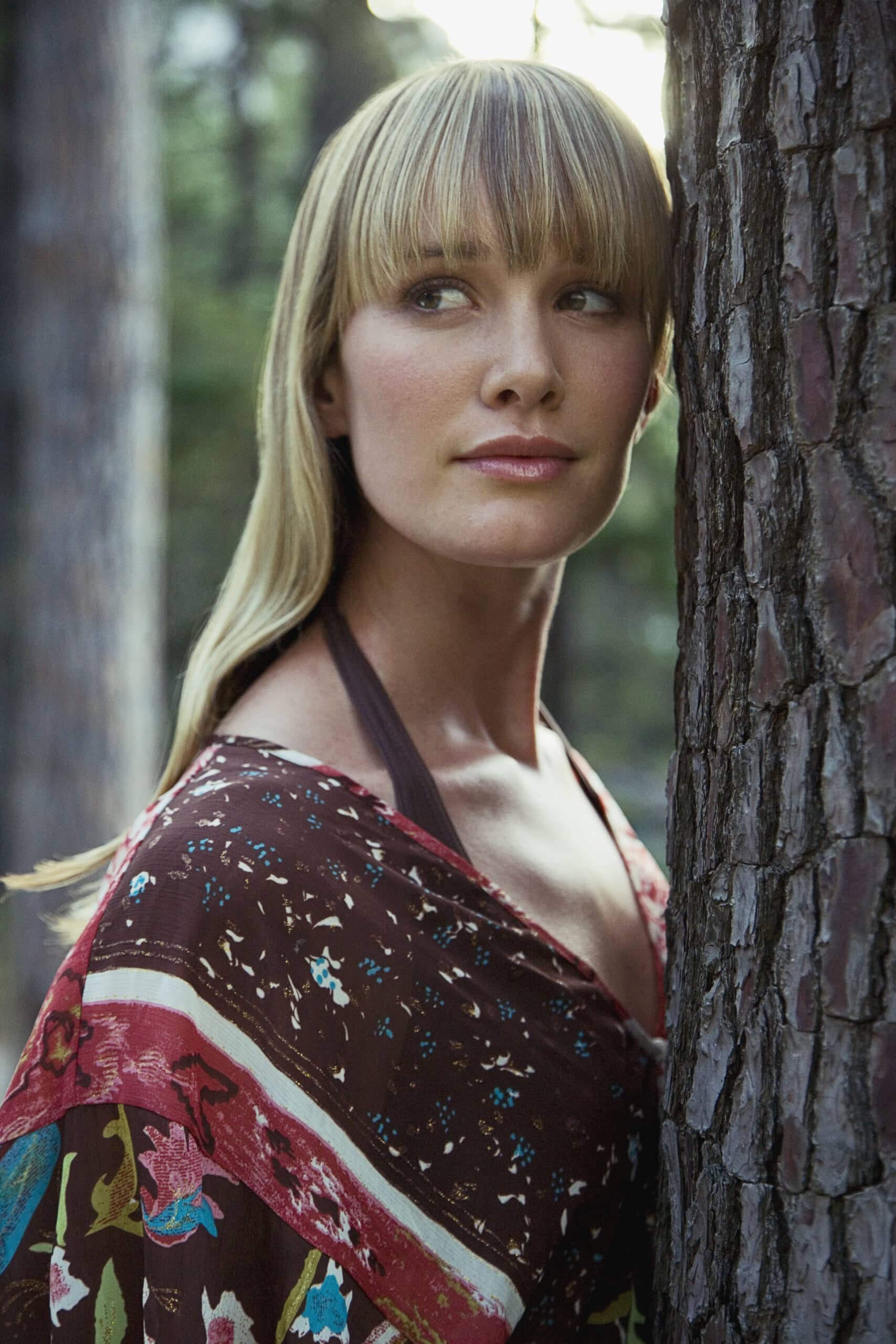 Eine Frau mit glattem blondem Haar und Pony steht dicht neben einem Baum. Sie trägt ein buntes, gemustertes Kleidungsstück mit Blumenmuster. Ihr Blick ist leicht zur Seite gerichtet, im Hintergrund ist ein Wald zu sehen, durch dessen Bäume sanftes Sonnenlicht fällt. © Fotografie Tomas Rodriguez