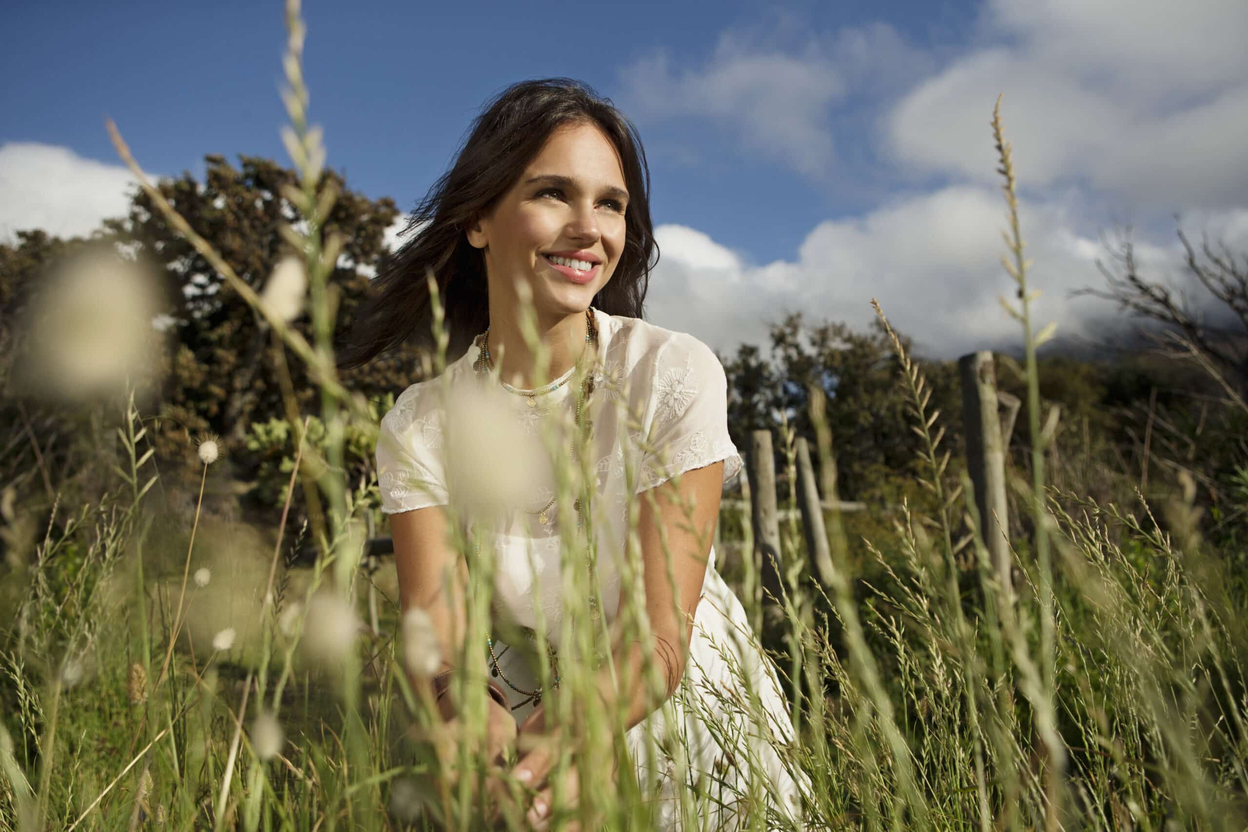 Eine Frau mit langem dunklem Haar und einem weißen Kleid kniet in einem Feld mit hohem Gras und lächelt vor der Kulisse von Bäumen und einem bewölkten blauen Himmel. © Fotografie Tomas Rodriguez