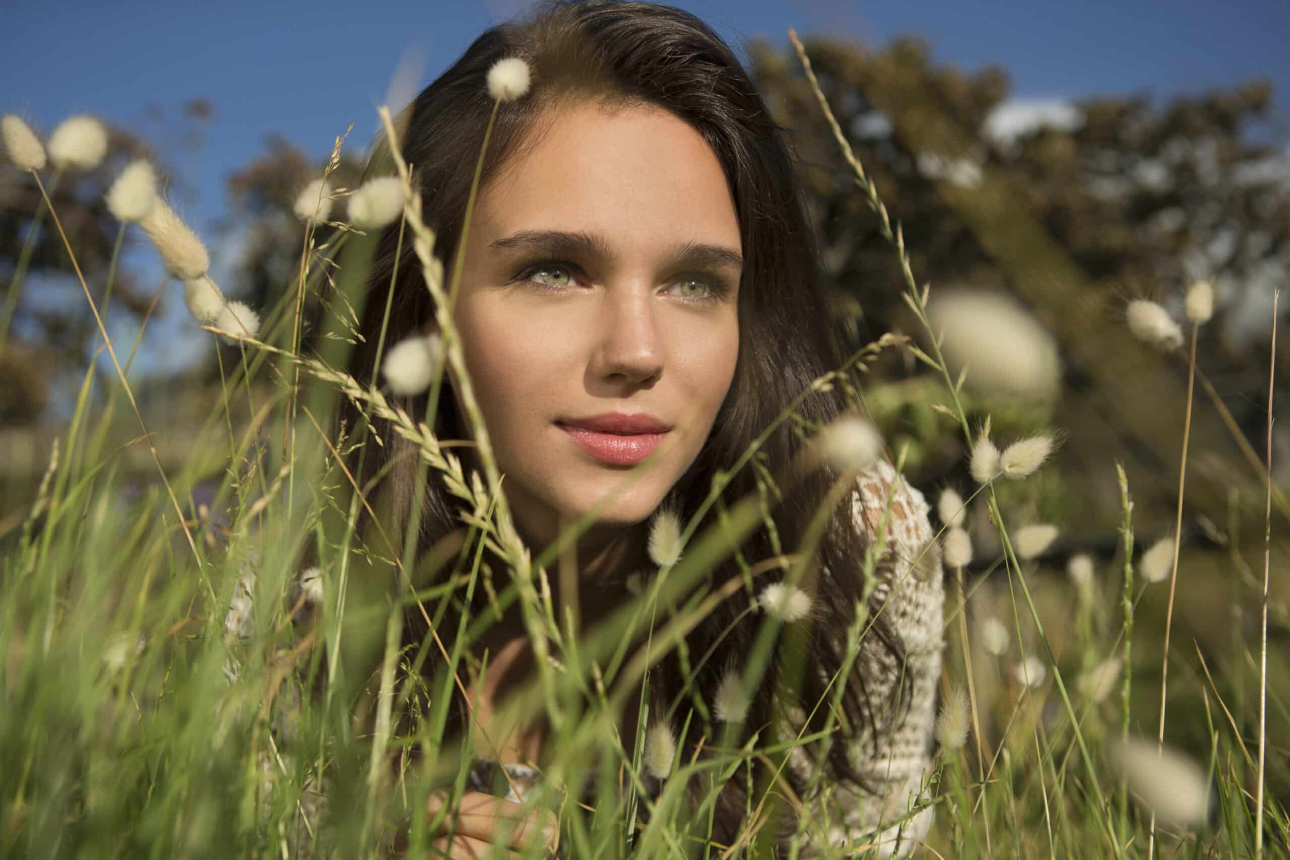 Eine junge Frau mit langen braunen Haaren und grünen Augen liegt in einem Feld mit hohem Gras und Wildblumen und blickt in die Ferne. Sie trägt ein helles Oberteil und im Hintergrund sind ein klarer blauer Himmel und verschwommene Bäume zu sehen. © Fotografie Tomas Rodriguez