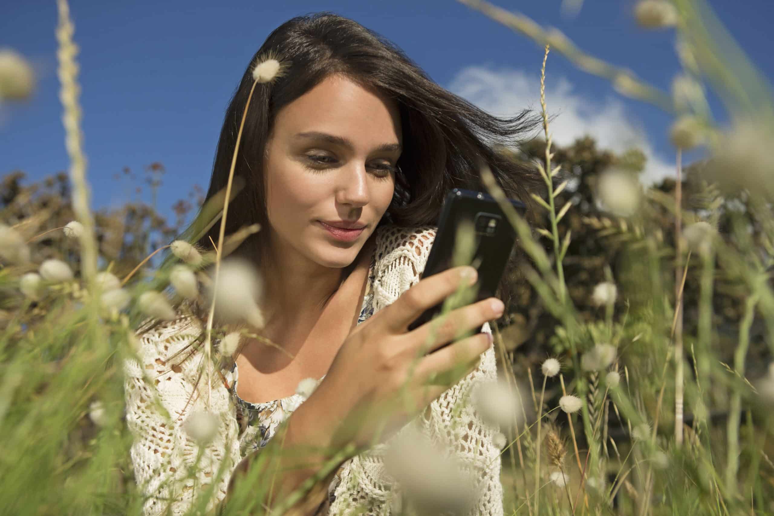 Eine Frau mit langen braunen Haaren sitzt in einem Feld voller Wildblumen und schaut auf ihr Smartphone. Sie trägt eine helle Strickjacke. Im Hintergrund ist der Himmel klar und blau, Bäume sind leicht zu erkennen. © Fotografie Tomas Rodriguez