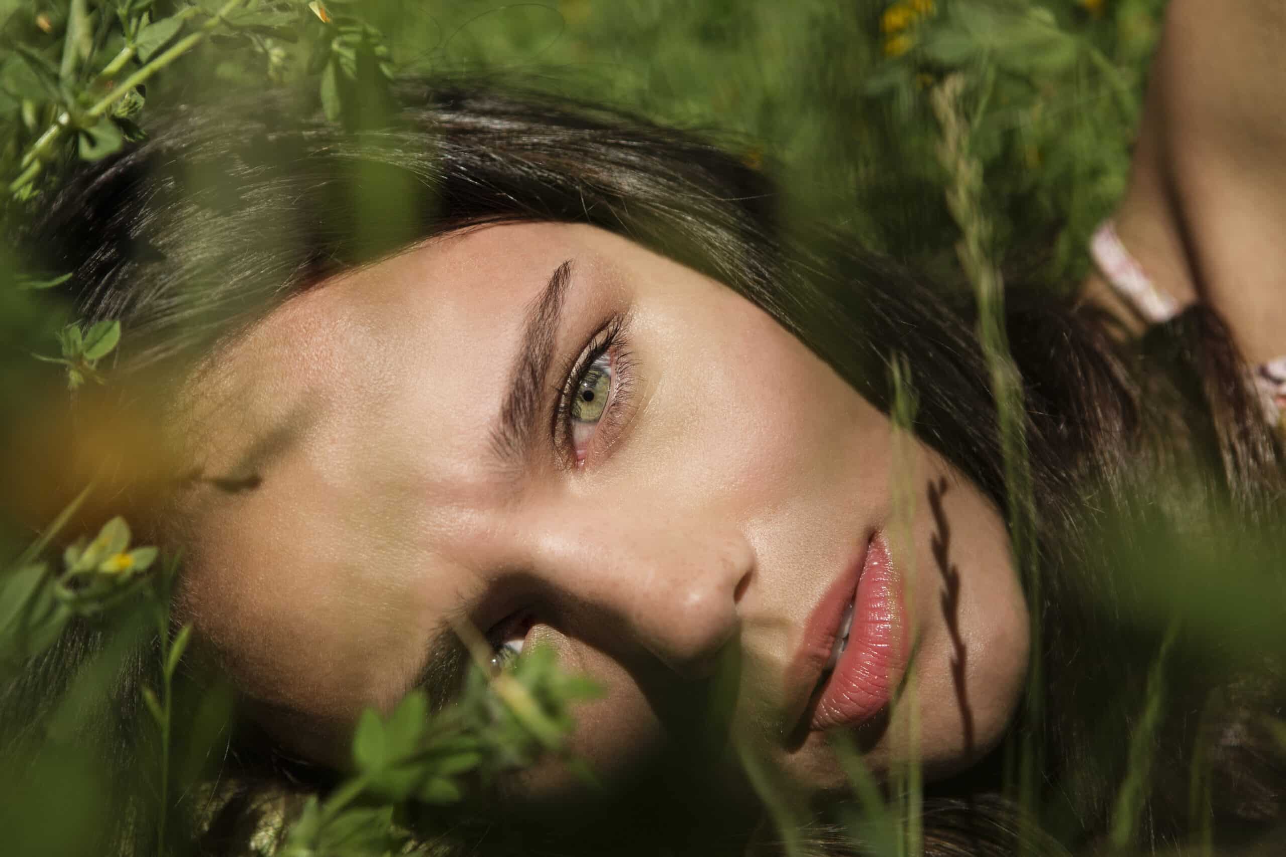 Eine Nahaufnahme des Gesichts einer Person, die auf grünem Gras liegt und einen heiteren Ausdruck zeigt. Die Person hat langes, dunkles Haar, grüne Augen und natürliches Make-up. Grashalme und kleine gelbe Blumen rahmen das Gesicht teilweise ein und schaffen eine friedliche, von der Natur inspirierte Szene. © Fotografie Tomas Rodriguez