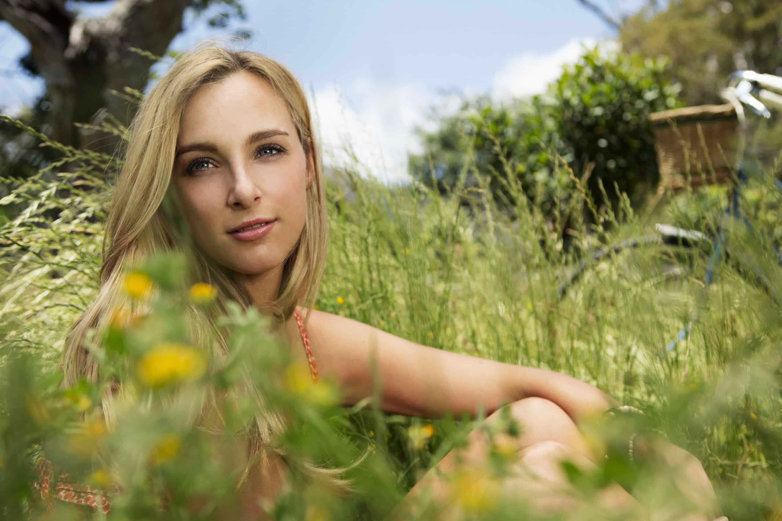 Eine Frau mit langen blonden Haaren sitzt auf einer Wiese, umgeben von hohem grünem Gras und gelben Blumen. Sie blickt mit sanftem Gesichtsausdruck direkt in die Kamera. Im Hintergrund sind ein Baum und ein verschwommenes Objekt in der Ferne zu sehen, vor einem hellen, sonnigen Himmel. © Fotografie Tomas Rodriguez