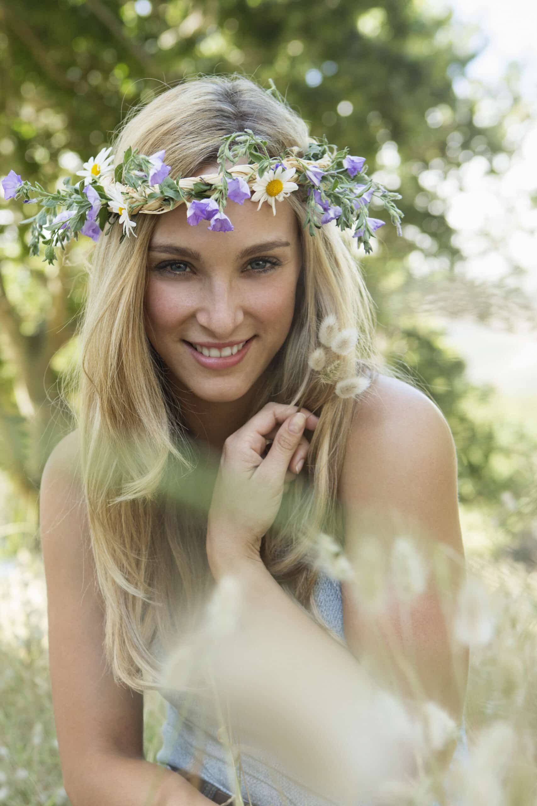 Eine Frau mit langen blonden Haaren und einem Blumenkranz aus violetten, gelben und weißen Blumen lächelt warm in die Kamera. Sie steht draußen auf einer Wiese mit verschwommenen grünen Bäumen im Hintergrund und berührt mit der Hand sanft ihr Haar. © Fotografie Tomas Rodriguez