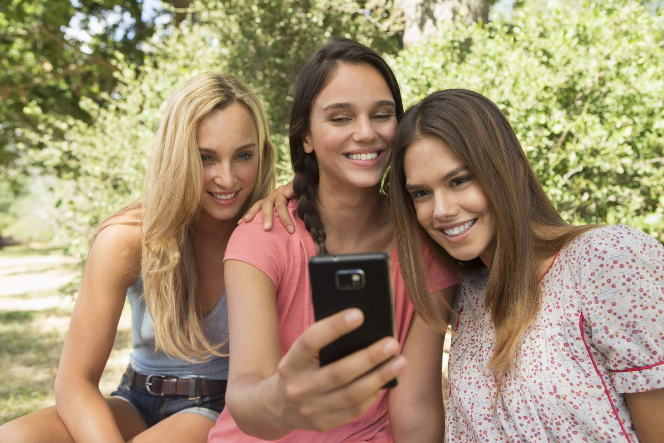 Drei Frauen sitzen dicht beieinander im Freien, lächeln und schauen auf ein Smartphone, das eine von ihnen hält. Sie scheinen ein Selfie zu machen. Der Hintergrund ist üppig und grün mit Bäumen und Laub. Alle wirken glücklich und entspannt. © Fotografie Tomas Rodriguez