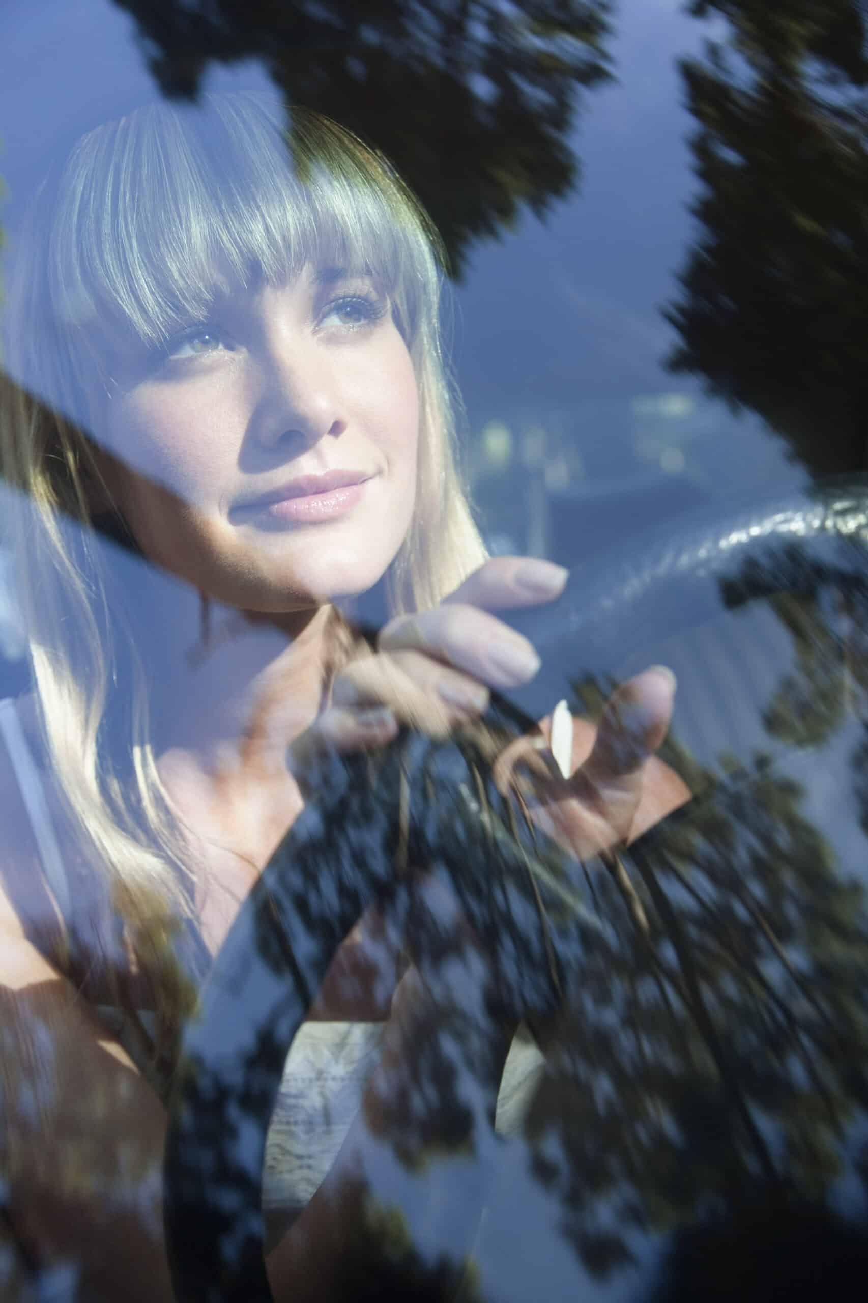 Eine Frau mit langen blonden Haaren blickt aus einer Windschutzscheibe und hält dabei das Lenkrad in der Hand. Das Sonnenlicht wirft Schatten von Ästen auf die Scheibe und überlagert so ihren nachdenklichen Gesichtsausdruck. © Fotografie Tomas Rodriguez