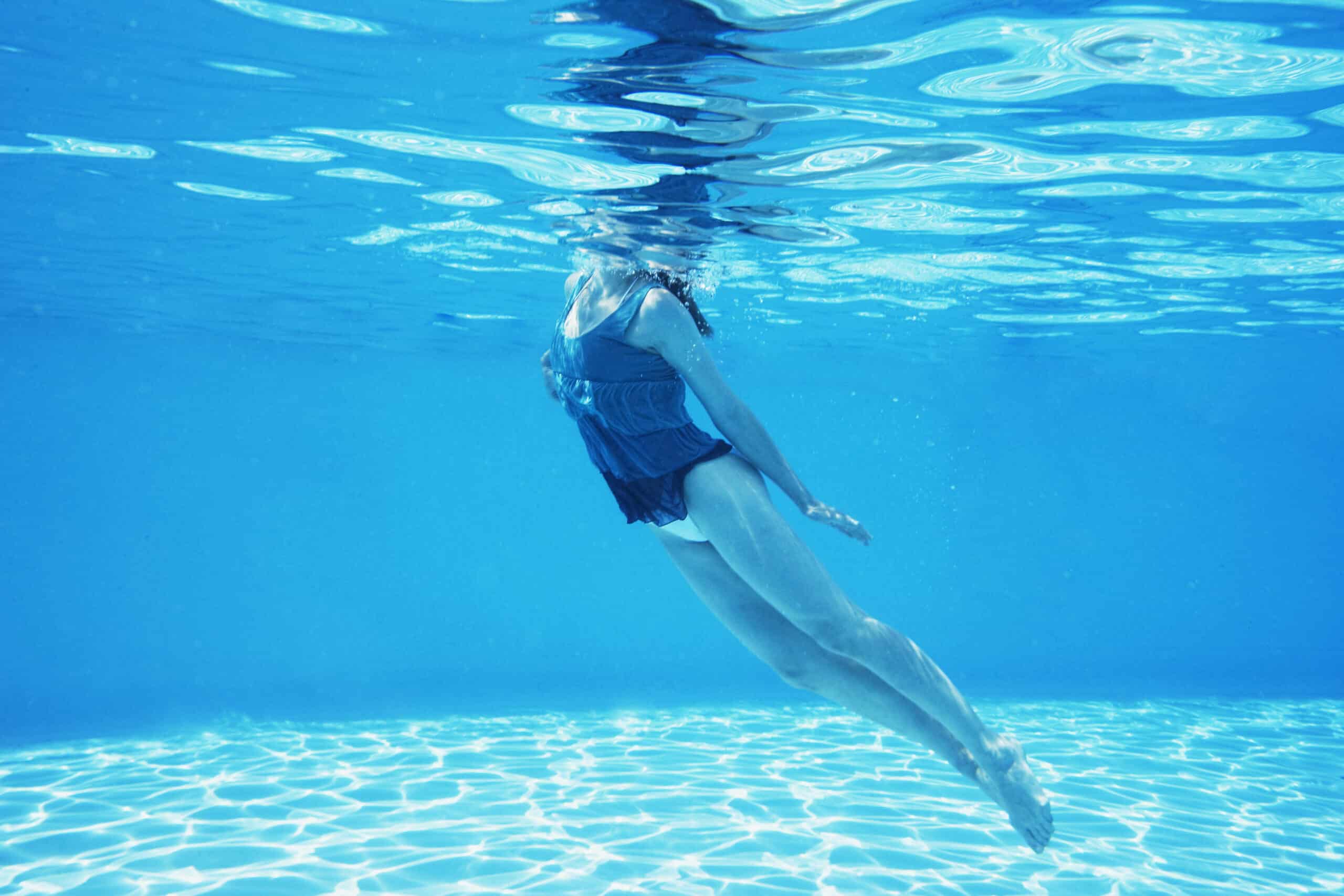 Eine Person schwimmt unter Wasser in einem klaren, blauen Pool. Sie trägt ein dunkles Oberteil und eine helle Hose. Das Wasser ist ruhig und das Sonnenlicht spiegelt sich im Poolboden. Der Schwimmer schwimmt horizontal nahe der Oberfläche, die Arme hängen an den Seiten. © Fotografie Tomas Rodriguez