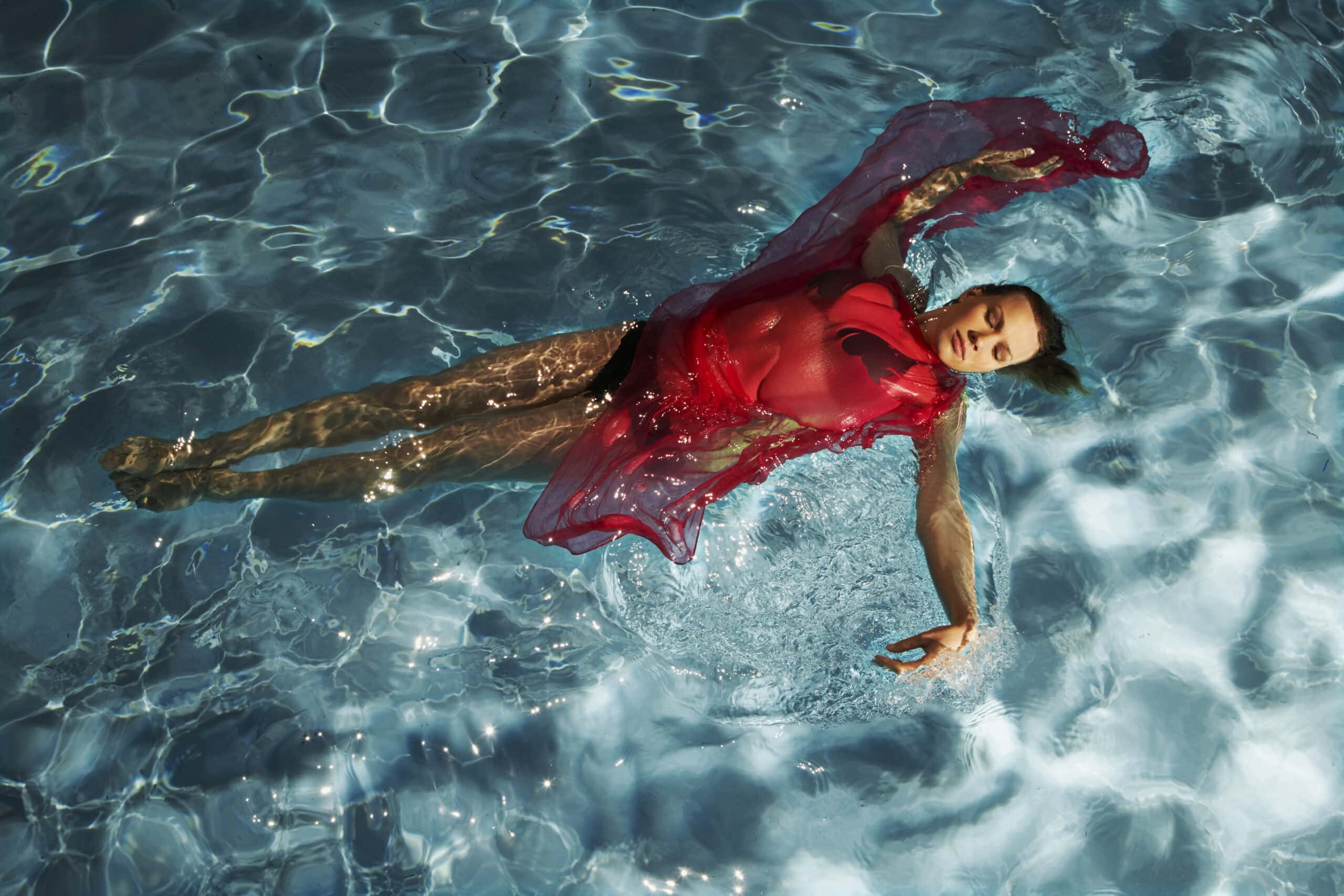 Eine Person in einem roten Kleid schwimmt auf dem Rücken in einem Schwimmbecken. Das Sonnenlicht fällt durch das Wasser und erzeugt einen schimmernden Effekt. Die Person wirkt entspannt, ein Arm ist ausgestreckt, der andere ruht auf dem Körper. © Fotografie Tomas Rodriguez