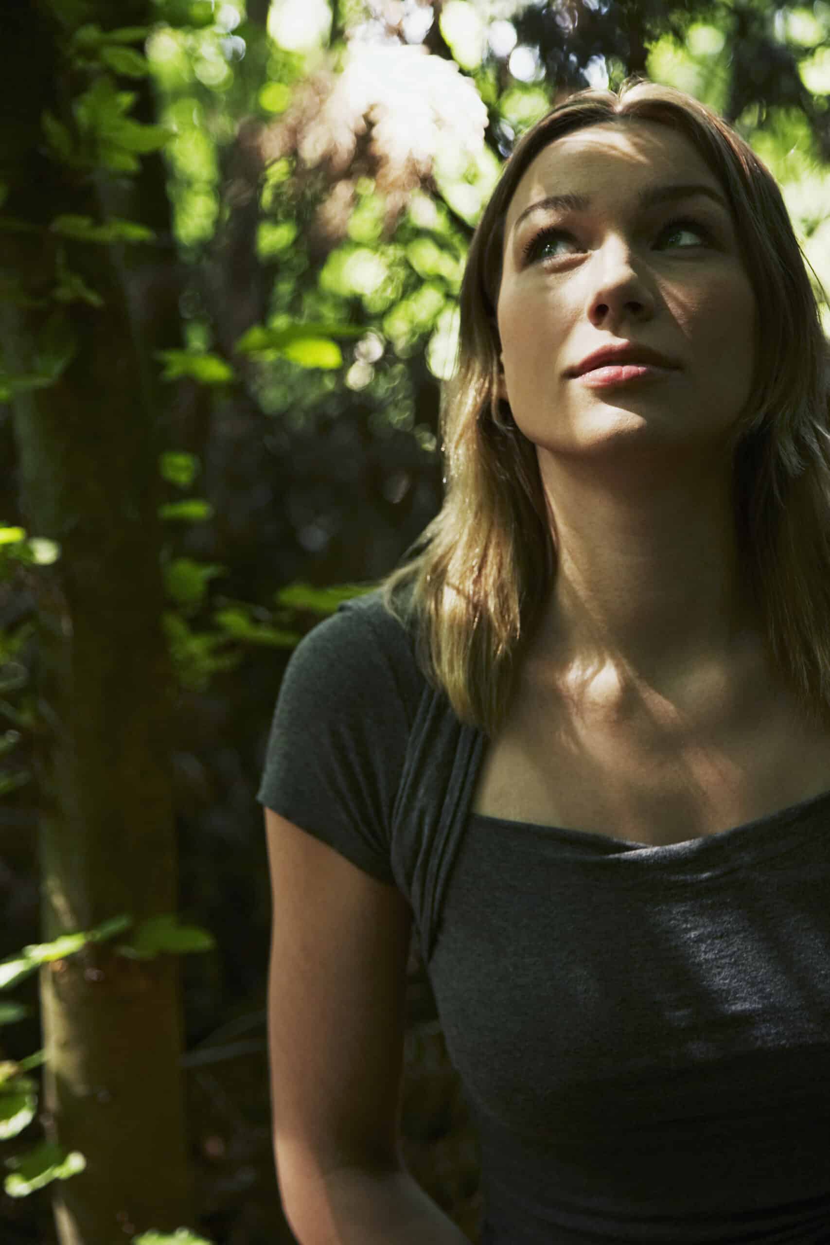 Eine Frau mit schulterlangem blondem Haar und einem dunkelgrauen T-Shirt steht in einem Wald. Sie blickt mit heiterem Gesichtsausdruck nach oben. Sonnenlicht fällt durch die Bäume und wirft gesprenkeltes Licht auf ihr Gesicht und die Umgebung. © Fotografie Tomas Rodriguez