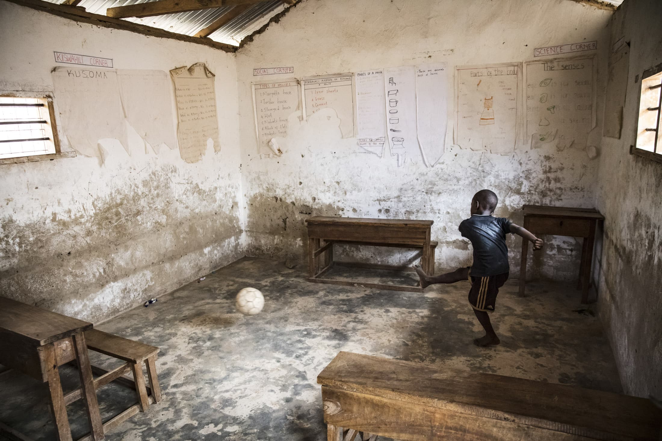 In einem spärlich eingerichteten Klassenzimmer spielt ein Kind mit einem Fußball. Der Raum hat ein paar Holzbänke und Tische, an den rauen, verputzten Wänden hängen Lehrplakate. Sonnenlicht fällt durch ein kleines vergittertes Fenster auf der linken Seite. © Fotografie Tomas Rodriguez