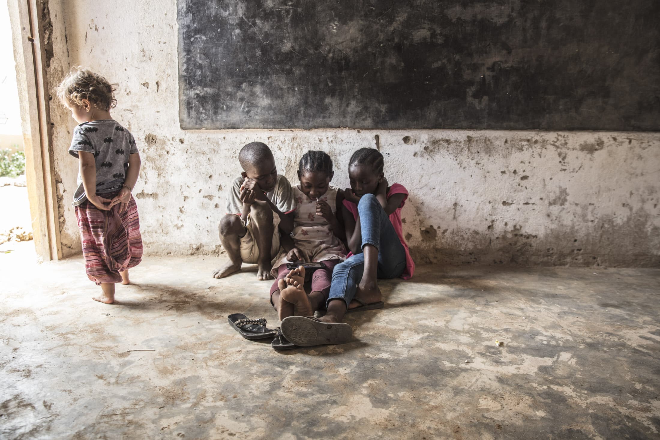 Fünf Kinder stehen drinnen vor einer strukturierten Wand, vier sitzen und sind mit einer gemeinsamen Aktivität beschäftigt, während eines daneben steht. Die sitzenden Kinder stehen eng beieinander, Schuhe liegen verstreut herum. Licht fällt durch eine Tür auf der linken Seite herein. © Fotografie Tomas Rodriguez