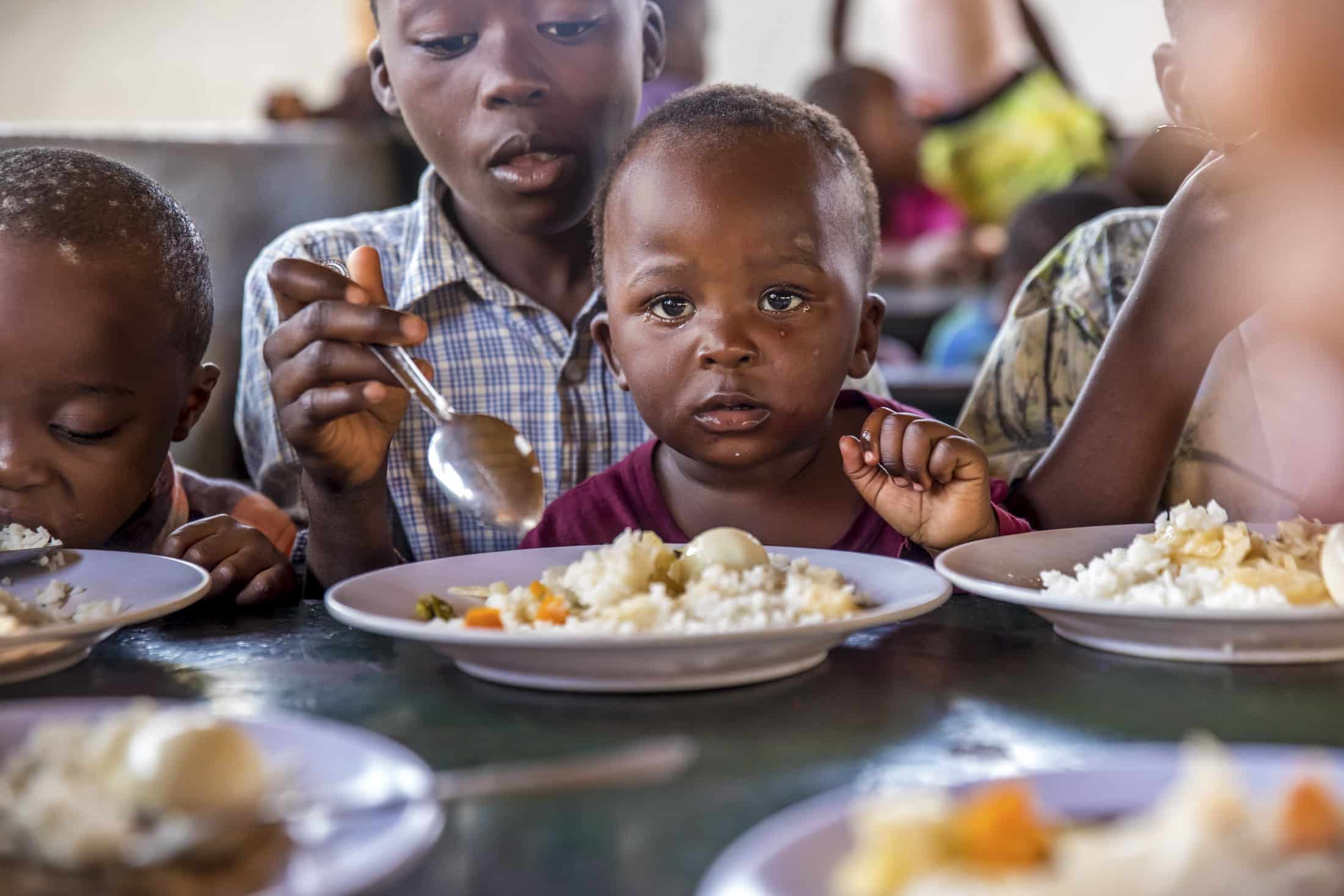 Kleine Kinder sitzen an einem Tisch mit Tellern voller Essen. Mehrere Kinder essen Reis mit Gemüse. Ein Kind starrt mit Essen in der Hand in die Kamera. Es scheint sich um eine gemeinsame Mahlzeit zu handeln. © Fotografie Tomas Rodriguez