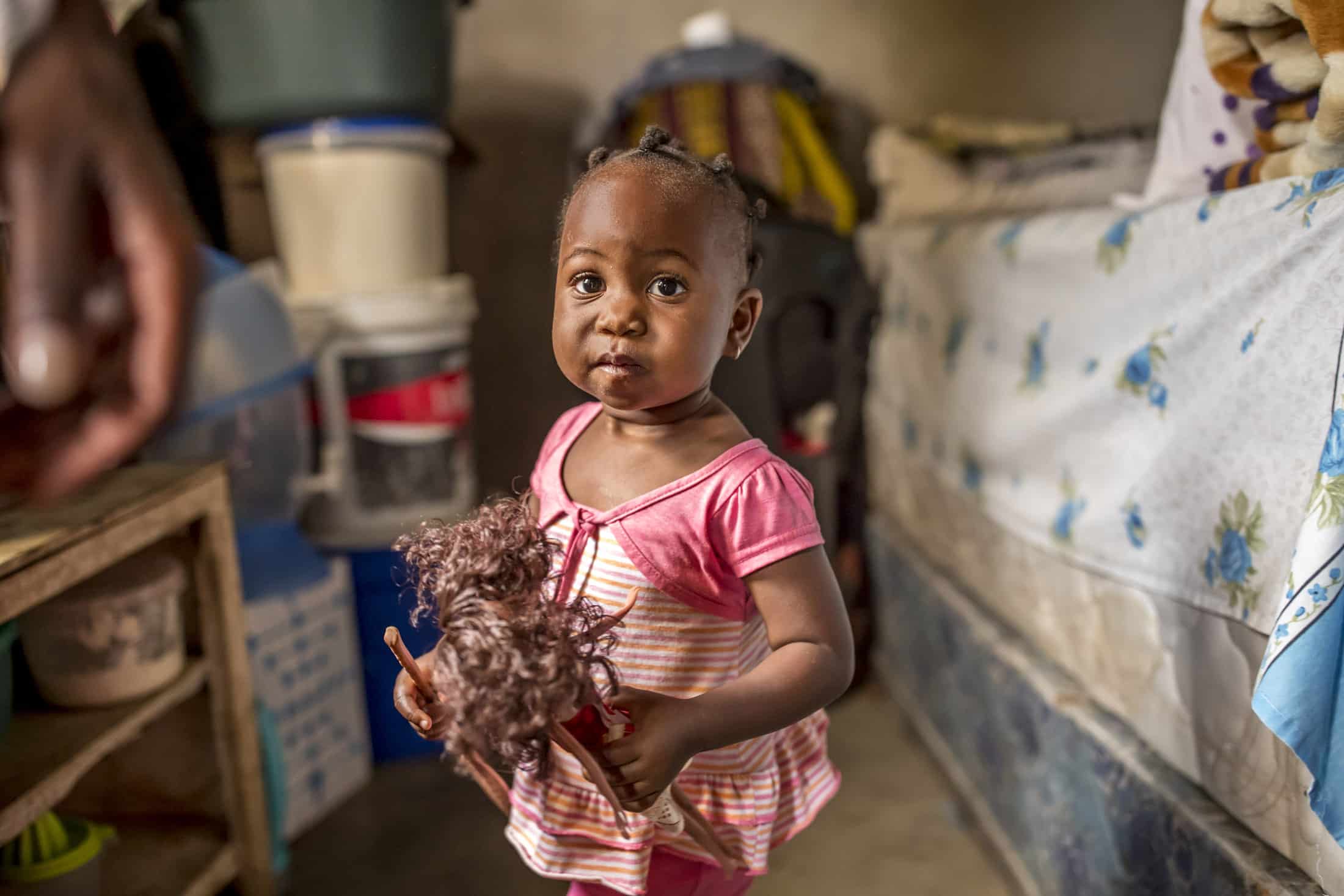 Ein kleines Kind mit geflochtenen Haaren steht in einem Zimmer und hält eine Puppe. Das Kind trägt ein gestreiftes Kleid mit rosa Ärmeln. Im Zimmer stehen verschiedene Gegenstände wie ein Bett mit geblümter Decke, Plastikbehälter und ein Regal. Das Kind blickt direkt in die Kamera. © Fotografie Tomas Rodriguez