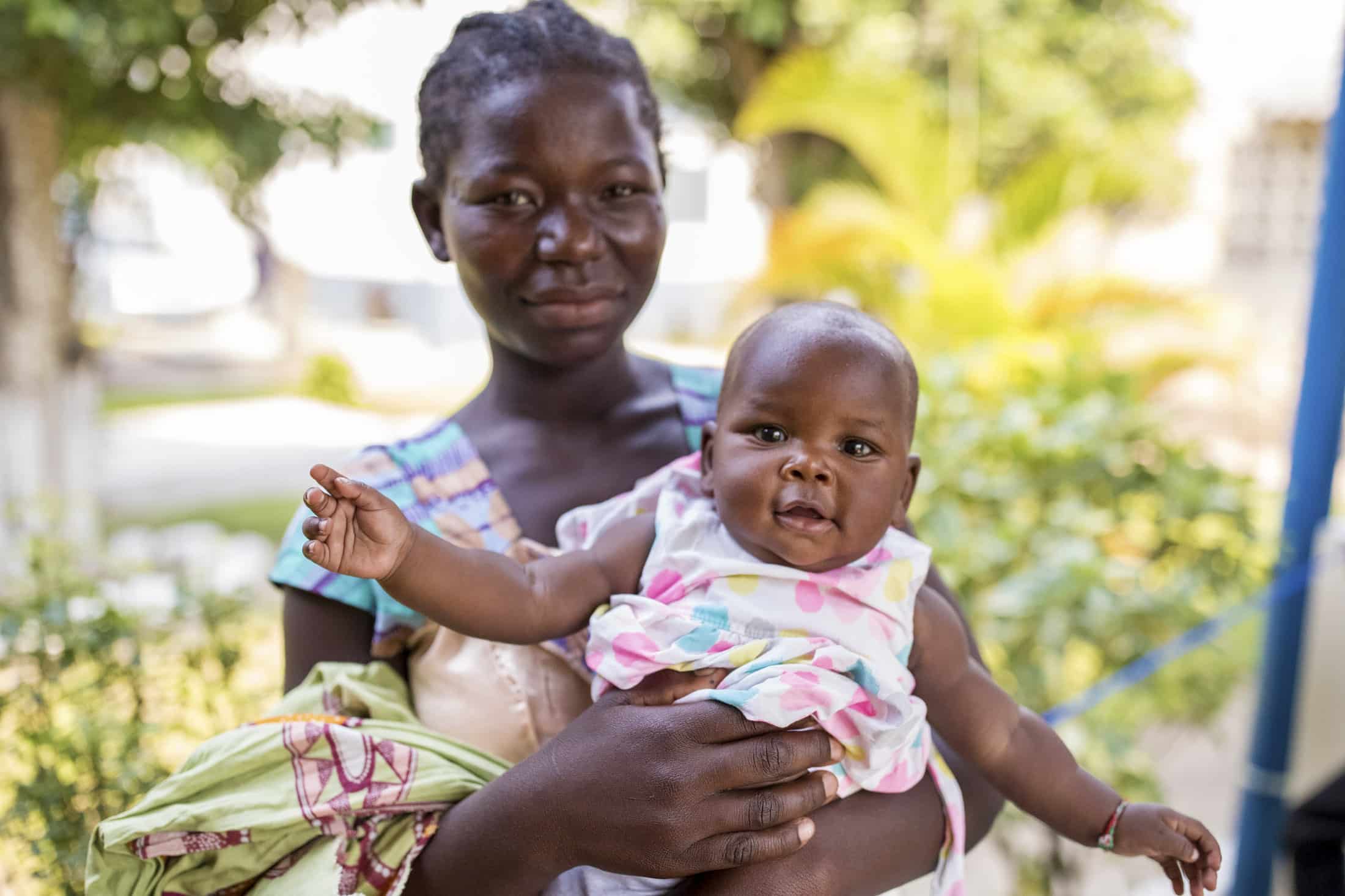 Eine Frau hält ein Baby im Arm, während sie draußen steht. Die Frau trägt ein gemustertes Kleid und hat einen ruhigen Gesichtsausdruck. Das Baby, das ein weißes Outfit mit bunten Mustern trägt, blickt mit offenem Mund und ausgestrecktem Arm in die Kamera. © Fotografie Tomas Rodriguez