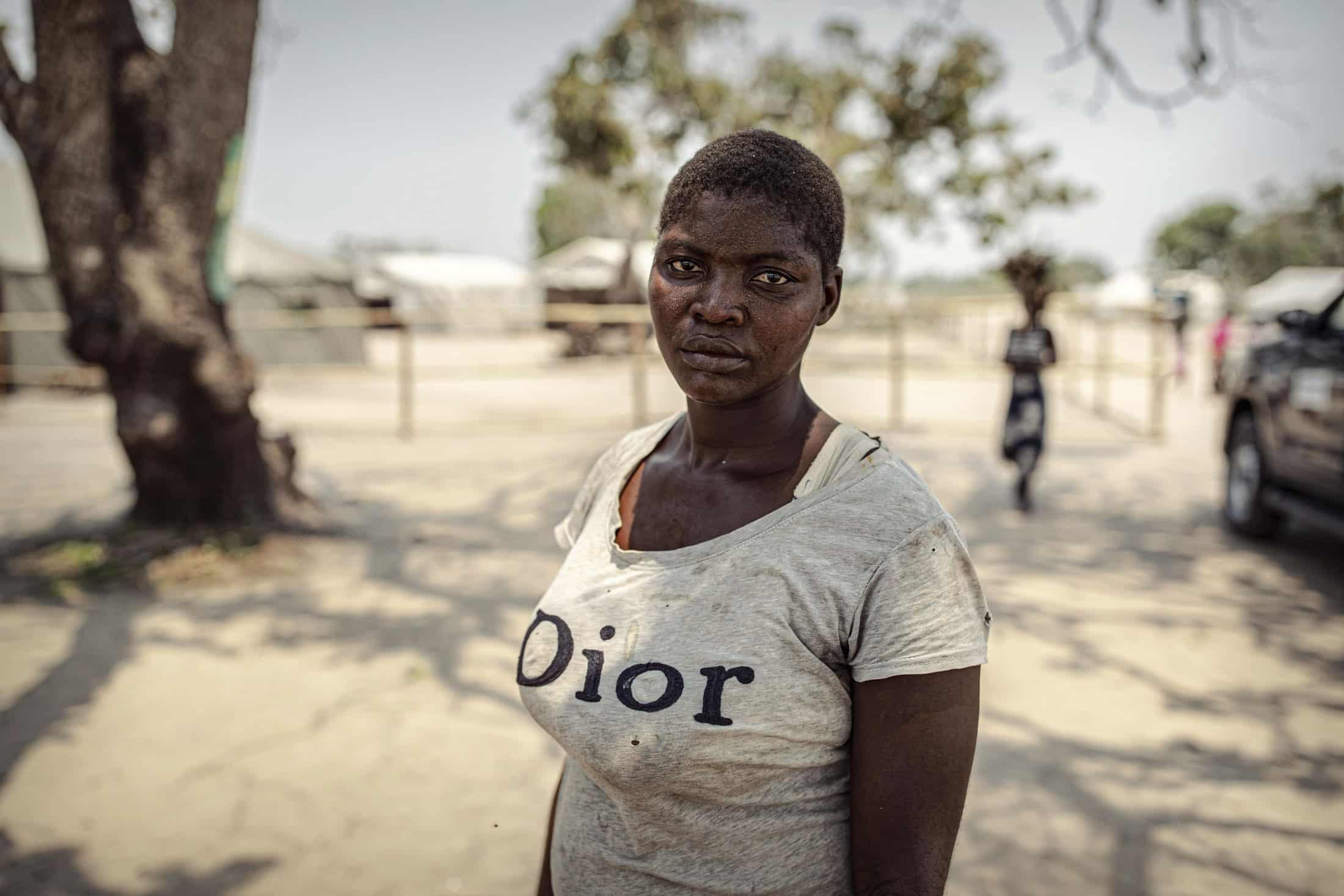 Eine Person in einem grauen Dior-T-Shirt steht mit ernster Miene im Schatten von Bäumen. Im Hintergrund sind verschwommene Figuren und Strukturen zu sehen, darunter ein Fahrzeug, was auf eine ländliche oder halbländliche Umgebung schließen lässt. Der Himmel erscheint klar und sonnig. © Fotografie Tomas Rodriguez