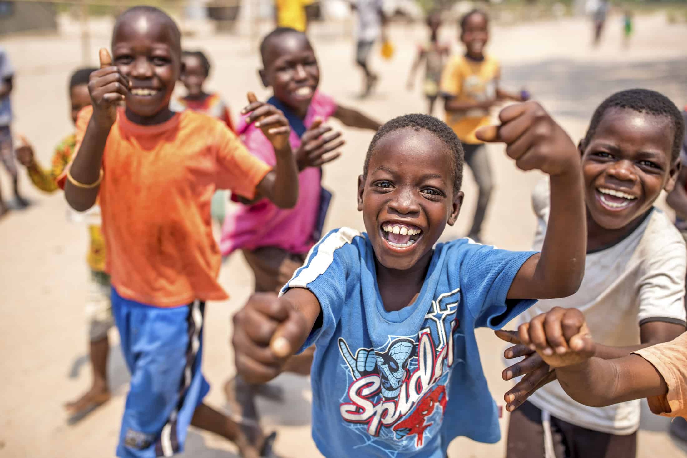 Eine Gruppe lächelnder Kinder im Freien, die aufgeregt mit ausgestreckten Armen auf die Kamera zulaufen. Der Junge im Vordergrund trägt ein blaues Spider-Man-Shirt und die anderen sind in bunte Kleidung gekleidet. Im Hintergrund ist eine verschwommene, sonnenbeschienene Szene im Freien zu sehen. © Fotografie Tomas Rodriguez