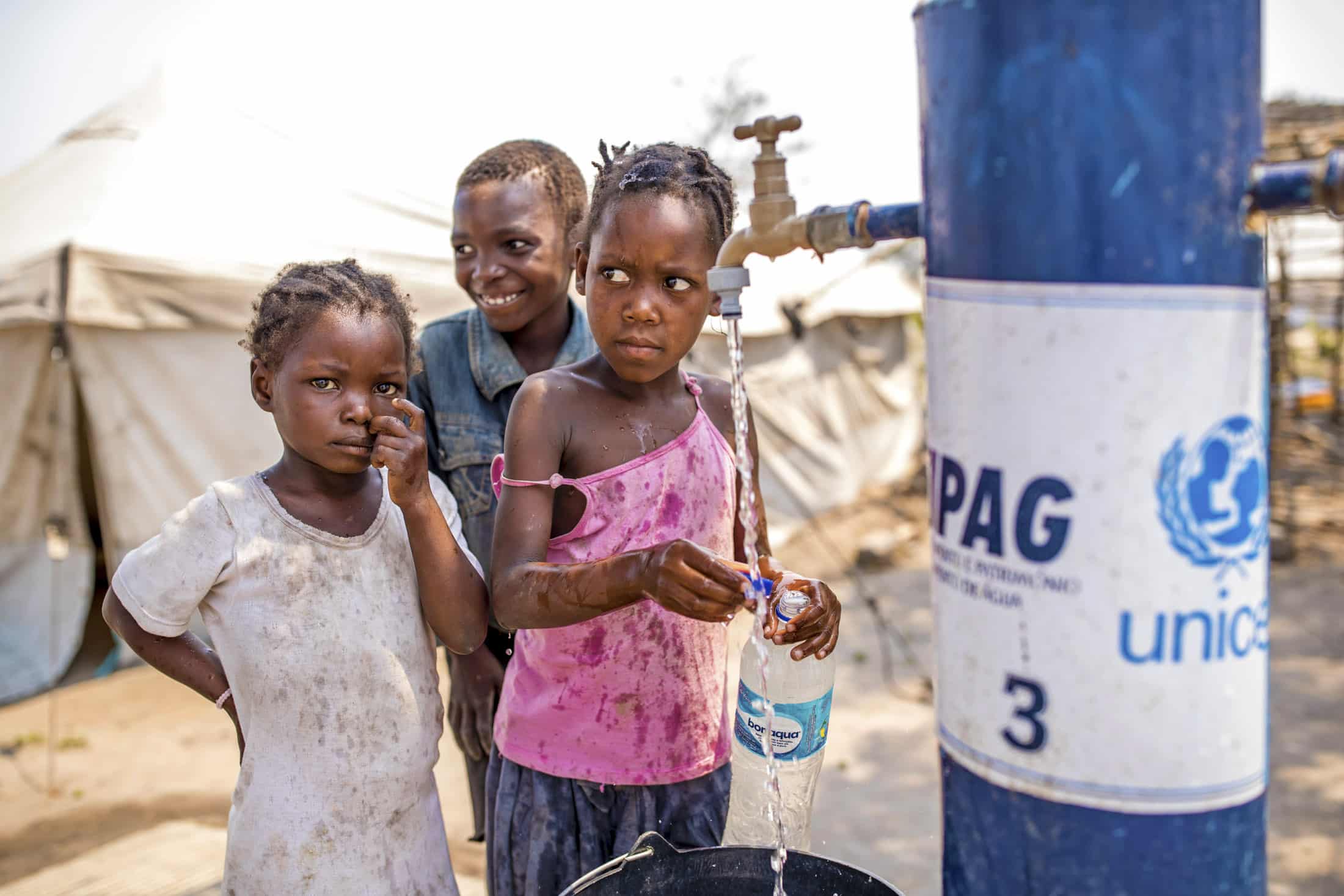 Drei Kinder versammeln sich um einen Wasserhahn. Eines füllt eine Flasche, ein anderes lächelt im Hintergrund und das dritte Kind schaut direkt in die Kamera. Im Hintergrund ist ein Zelt zu sehen. Auf dem Wasserhahn sind die Aufschriften „UNICEF“ und „IPAG“ zu sehen. Es scheint sich um ein Hilfslager zu handeln. © Fotografie Tomas Rodriguez