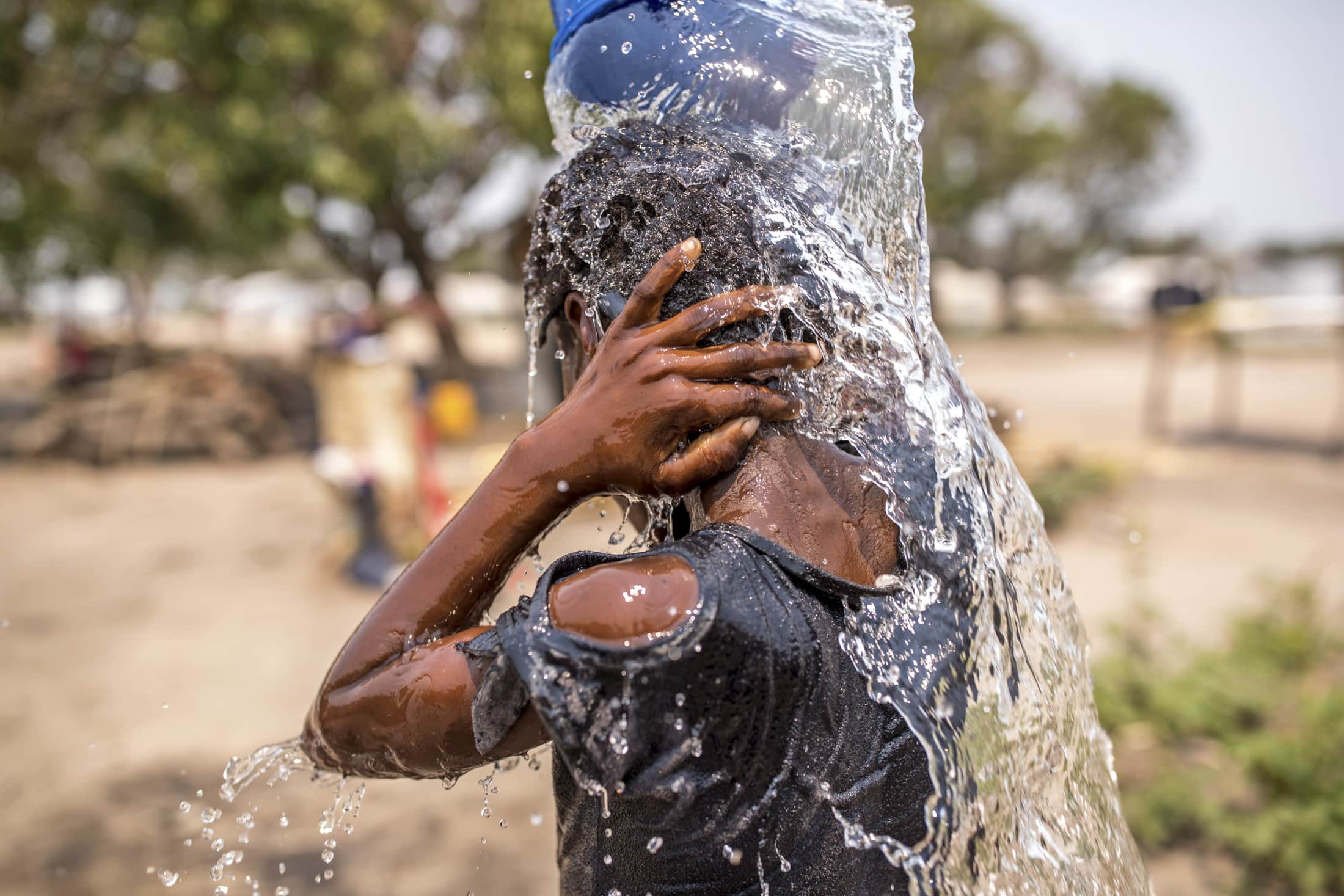 Ein junger Mann gießt sich aus einem blauen Behälter Wasser über den Kopf. Er trägt ein schwarzes Hemd mit Löchern. Das Bild fängt den Moment ein, in dem das Wasser herunterspritzt. Im Hintergrund ist eine Außenkulisse mit verschwommenen Bäumen zu sehen. © Fotografie Tomas Rodriguez