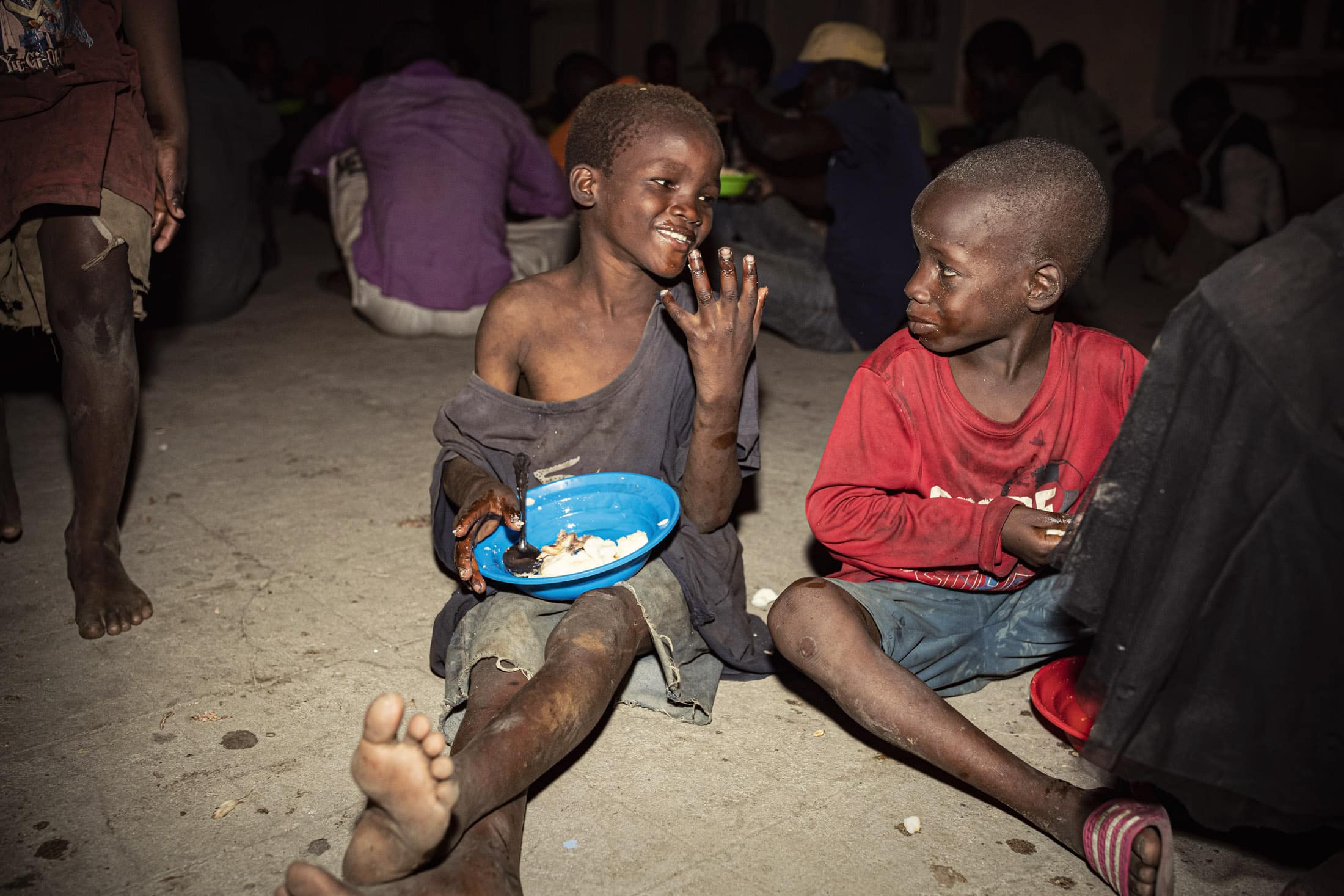 Zwei kleine Jungen sitzen auf dem Boden und essen aus blauen Plastikschüsseln. Ein Junge lacht und zeigt dem anderen seine Hand, der neugierig zusieht. Beide sind barfuß und tragen abgetragene Kleidung. Im Hintergrund sind Menschen zu sehen, die ebenfalls sitzen und essen. © Fotografie Tomas Rodriguez