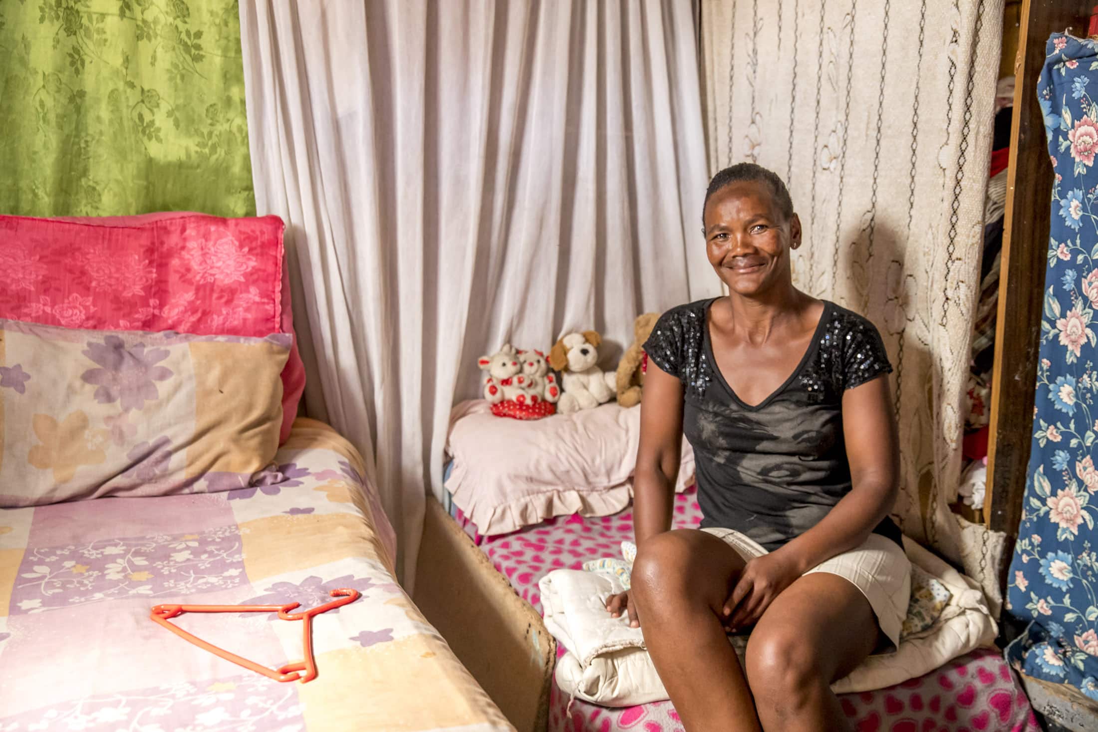 Eine Frau mit kurzen Haaren und schwarzem Hemd sitzt auf einem kleinen, ordentlich gemachten Bett in einem gemütlichen Zimmer mit bunten Tagesdecken, Stofftieren im Hintergrund und hängenden Vorhängen. Sie lächelt herzlich in die Kamera. © Fotografie Tomas Rodriguez