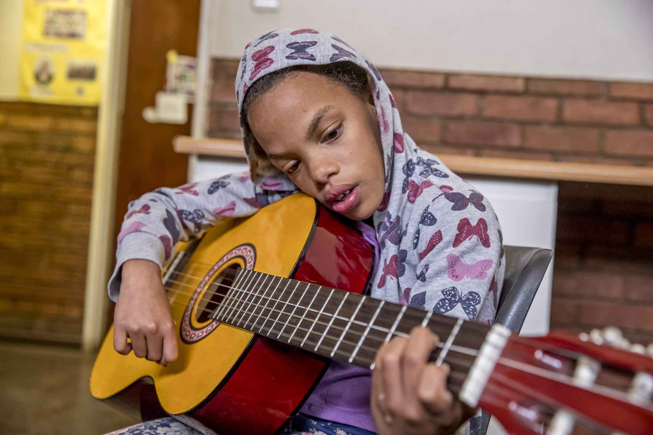 Eine Person trägt einen Kapuzenpullover und spielt konzentriert Akustikgitarre. Sie sitzt drinnen auf einem Stuhl. Im Hintergrund sind eine Ziegelwand und eine Holztür mit einigen Plakaten zu sehen. © Fotografie Tomas Rodriguez