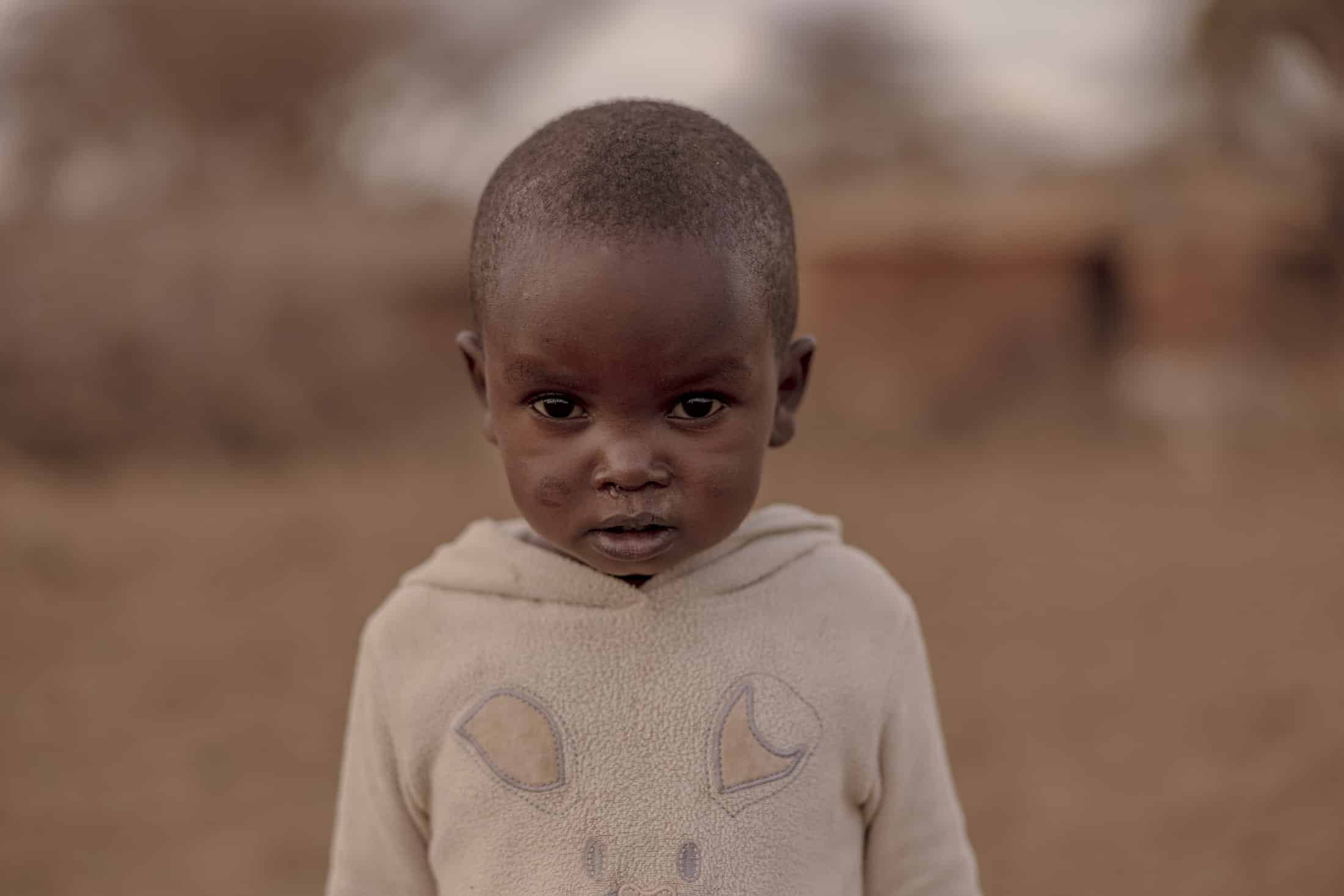 Ein kleines Kind steht im Freien und trägt einen hellen Kapuzenpullover mit einfachen Mustern auf der Vorderseite. Der Gesichtsausdruck des Kindes ist ernst und der Hintergrund ist mit erdigen Tönen und undeutlichen Formen sanft verschwommen. © Fotografie Tomas Rodriguez