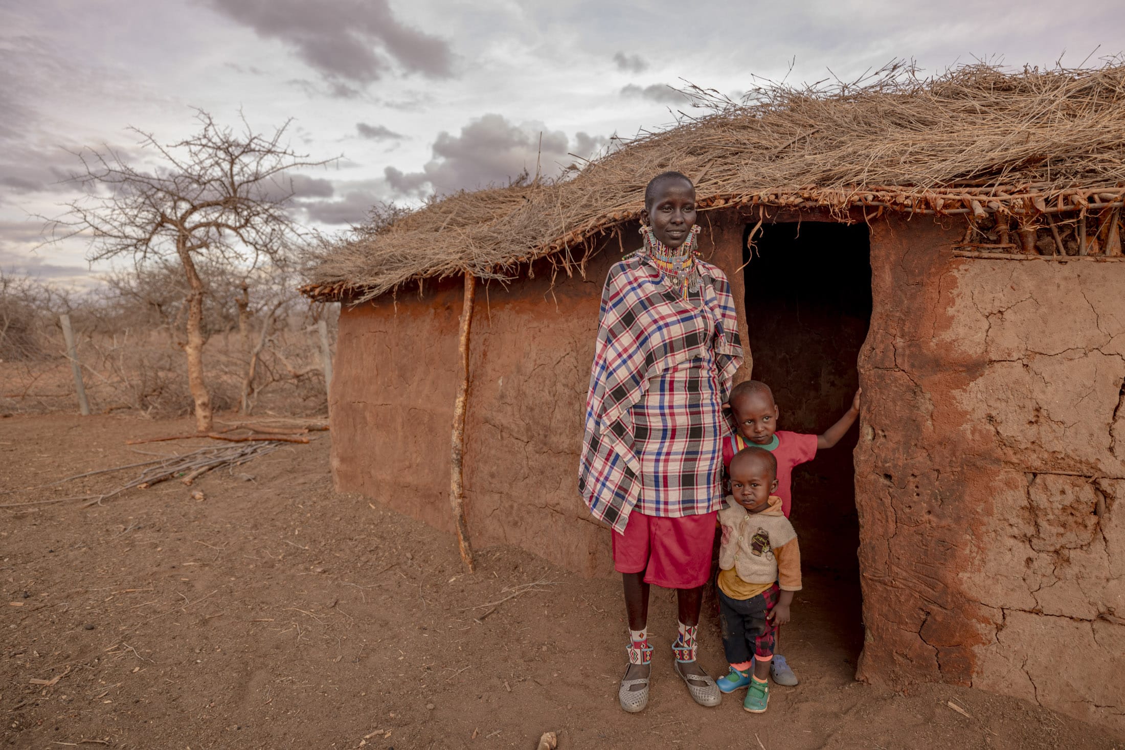 Eine Person in traditioneller Kleidung steht mit zwei kleinen Kindern vor einer Lehmhütte mit Strohdach. Die Person und ein Kind posieren vor der Tür, während das andere Kind hinter der Tür hervorlugt. Die Landschaft ist trocken mit spärlichen, blattlosen Bäumen. © Fotografie Tomas Rodriguez