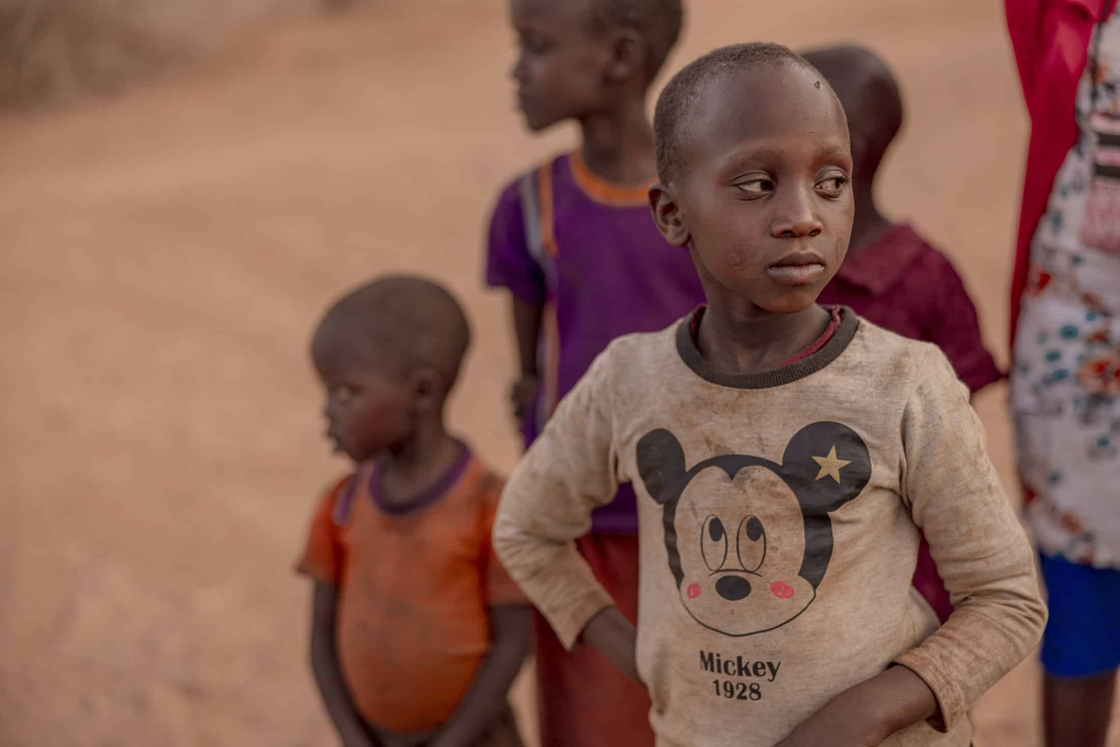 Ein kleines Kind in einem Mickey-Mouse-Shirt steht mit neugierigem Gesichtsausdruck im Freien. Im Hintergrund sind mehrere andere Kinder zu sehen, eines davon blickt zur Seite und trägt ein orangefarbenes Shirt. Die Landschaft wirkt staubig und warm, in gedämpften Erdtönen. © Fotografie Tomas Rodriguez