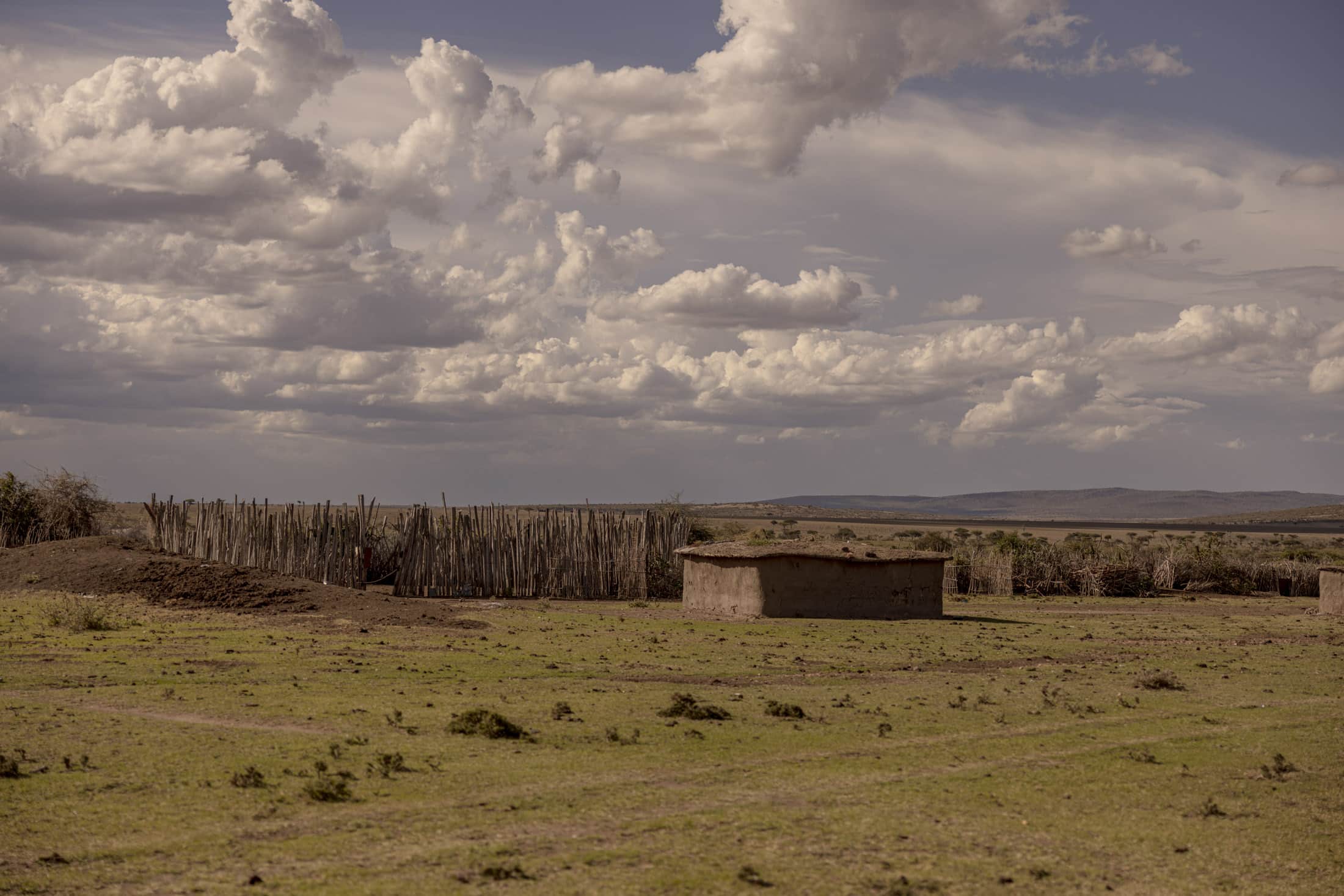 Eine weite Landschaft mit einer einfachen runden Erdstruktur im Vordergrund, umgeben von grünem Gras. Links umschließt ein Holzzaun ein Gebiet. Der Himmel ist mit großen, flauschigen Wolken gefüllt. In der Ferne sind flaches Gelände und eine Bergkette sichtbar. © Fotografie Tomas Rodriguez