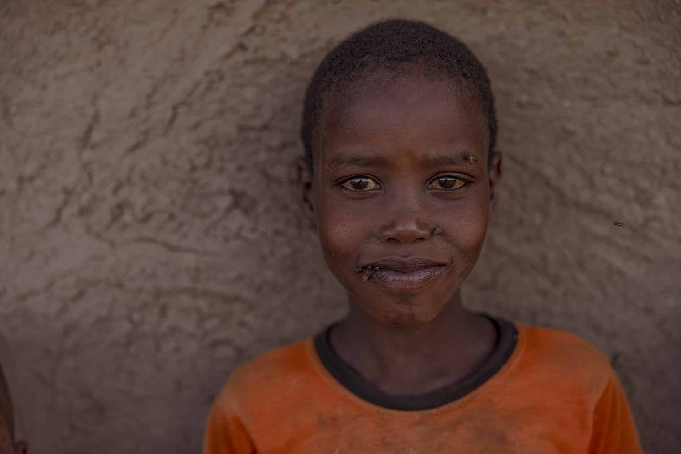 Eine junge Person mit kurzen Haaren steht vor einer strukturierten, erdigen Wand. Sie trägt ein orangefarbenes Hemd und hat einen ruhigen Gesichtsausdruck. Sie lächelt sanft in die Kamera. Ihr Gesicht ist dezent beleuchtet, wodurch ihre Gesichtszüge vor dem gedämpften Hintergrund hervorgehoben werden. © Fotografie Tomas Rodriguez