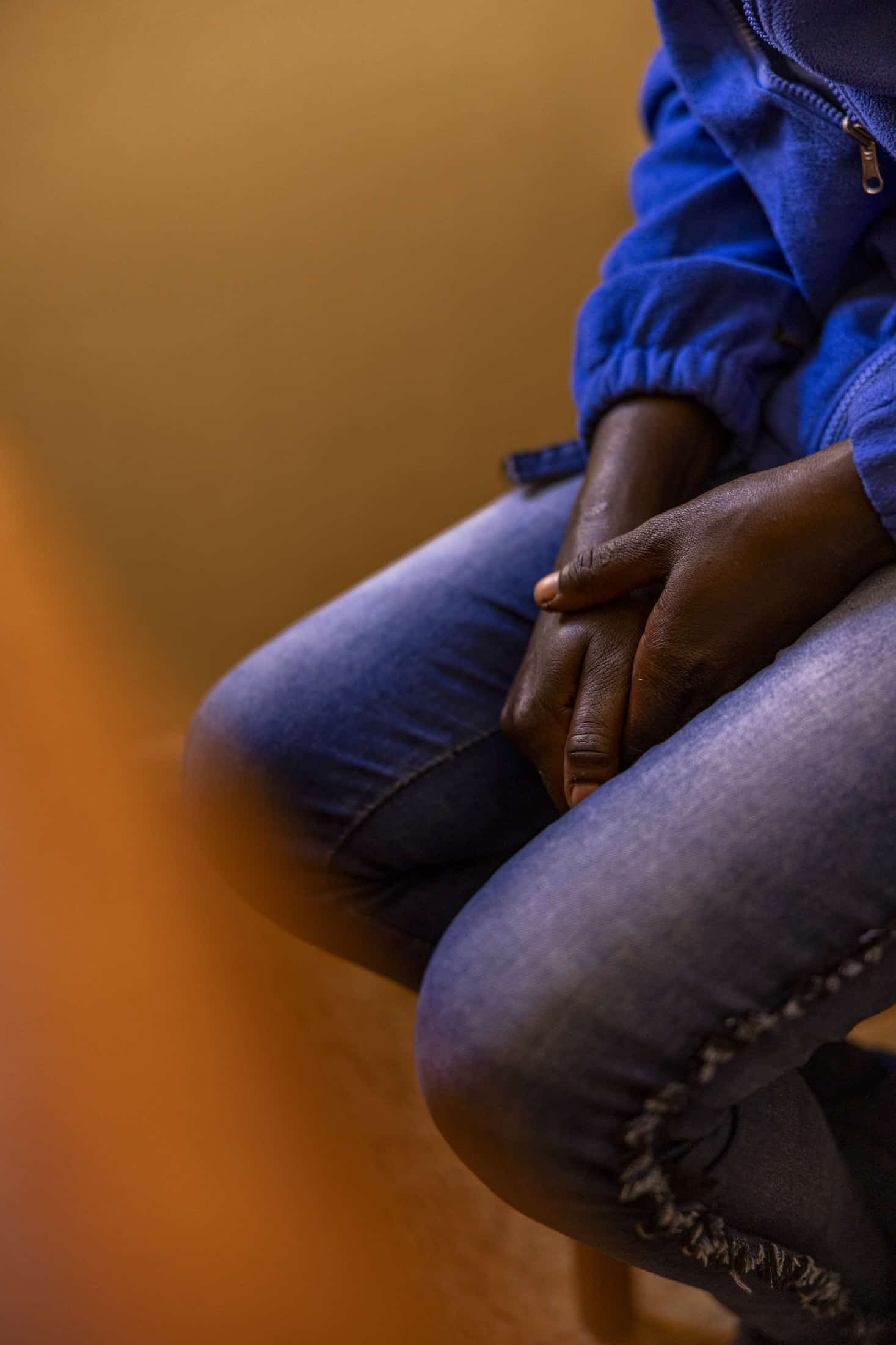 Eine Person in blauer Jacke und Jeans sitzt mit gefalteten Händen im Schoß. Der Hintergrund ist unscharf und weist eine warme, weiche Beleuchtung auf. © Fotografie Tomas Rodriguez